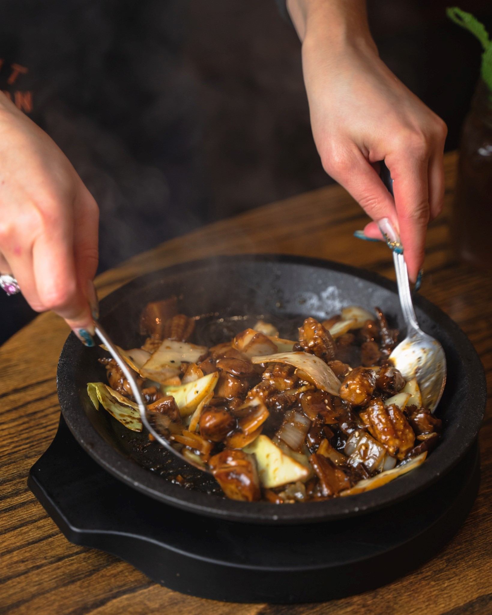Hands using chopsticks over a sizzling hot plate with vegetables and sauce.