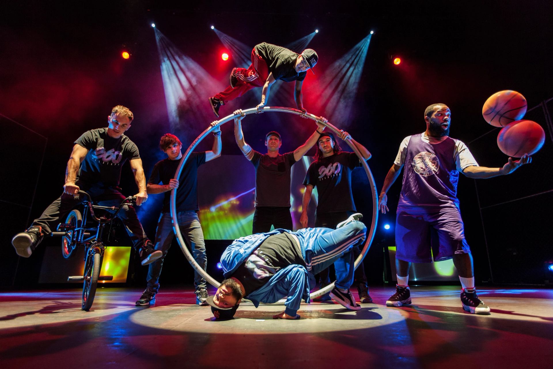 Street performers on stage doing BMX, breakdance and basketball tricks under colourful lights.