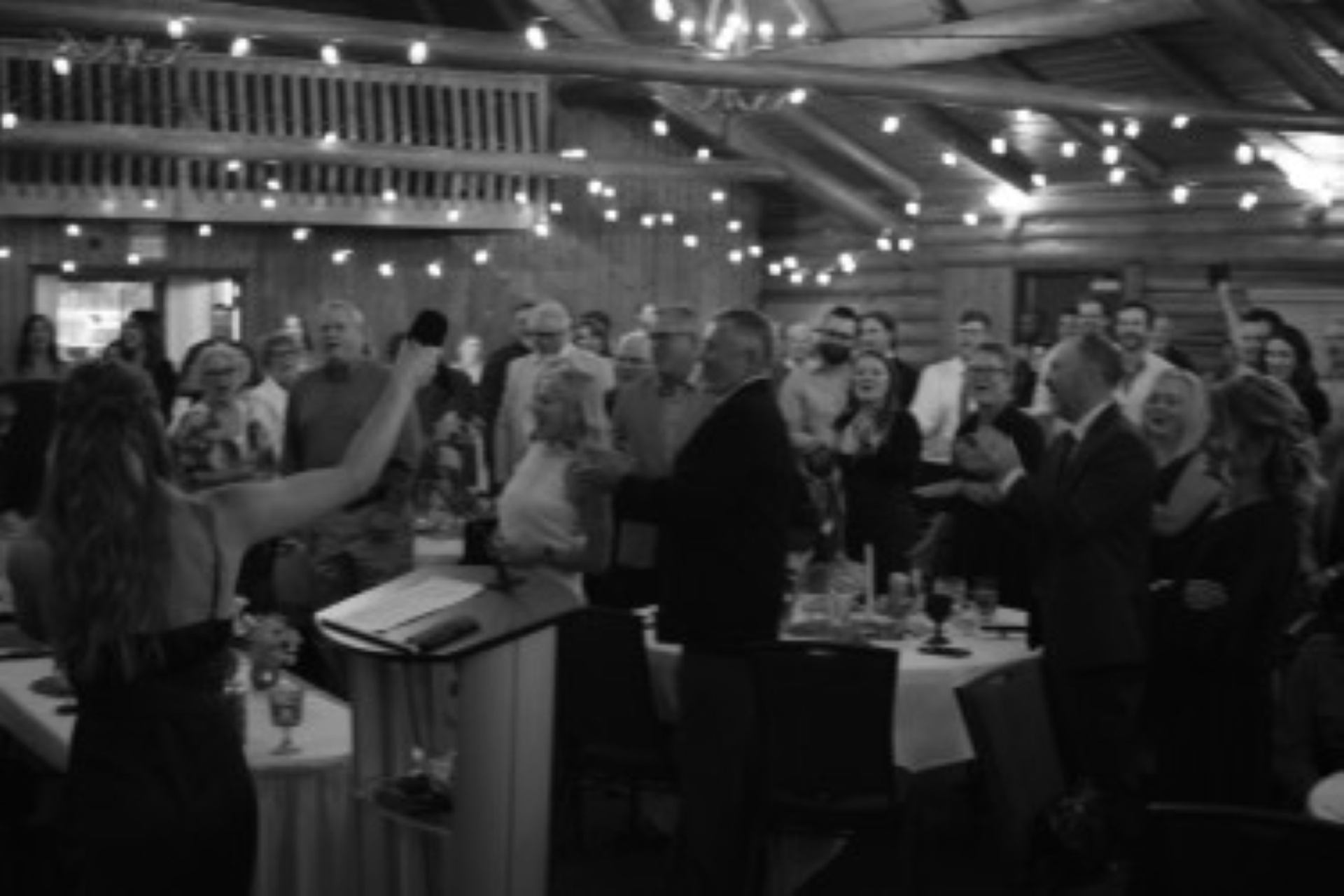 Crowd gathers around dining tables beneath string lights in a wooden hall.