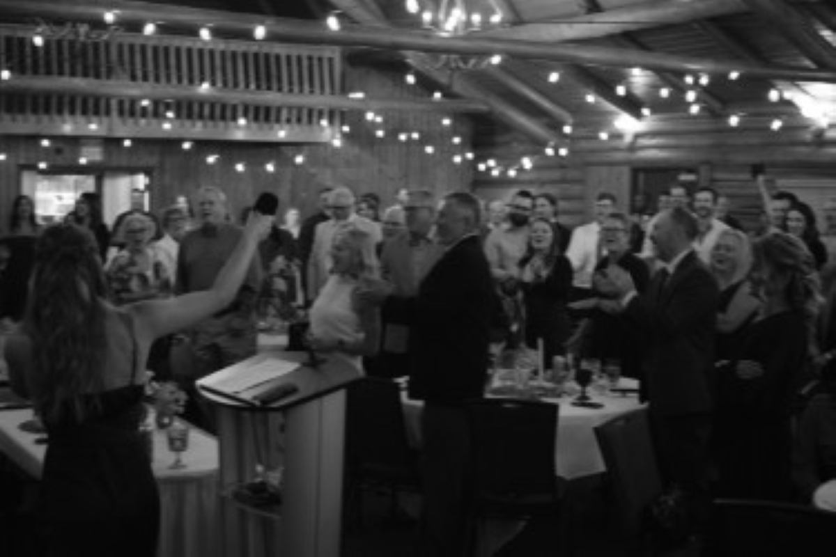 Crowd gathers around dining tables beneath string lights in a wooden hall.