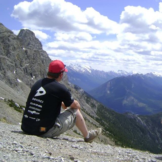 Person in red cap sits on rocky ridge overlooking mountain valley and clouds.