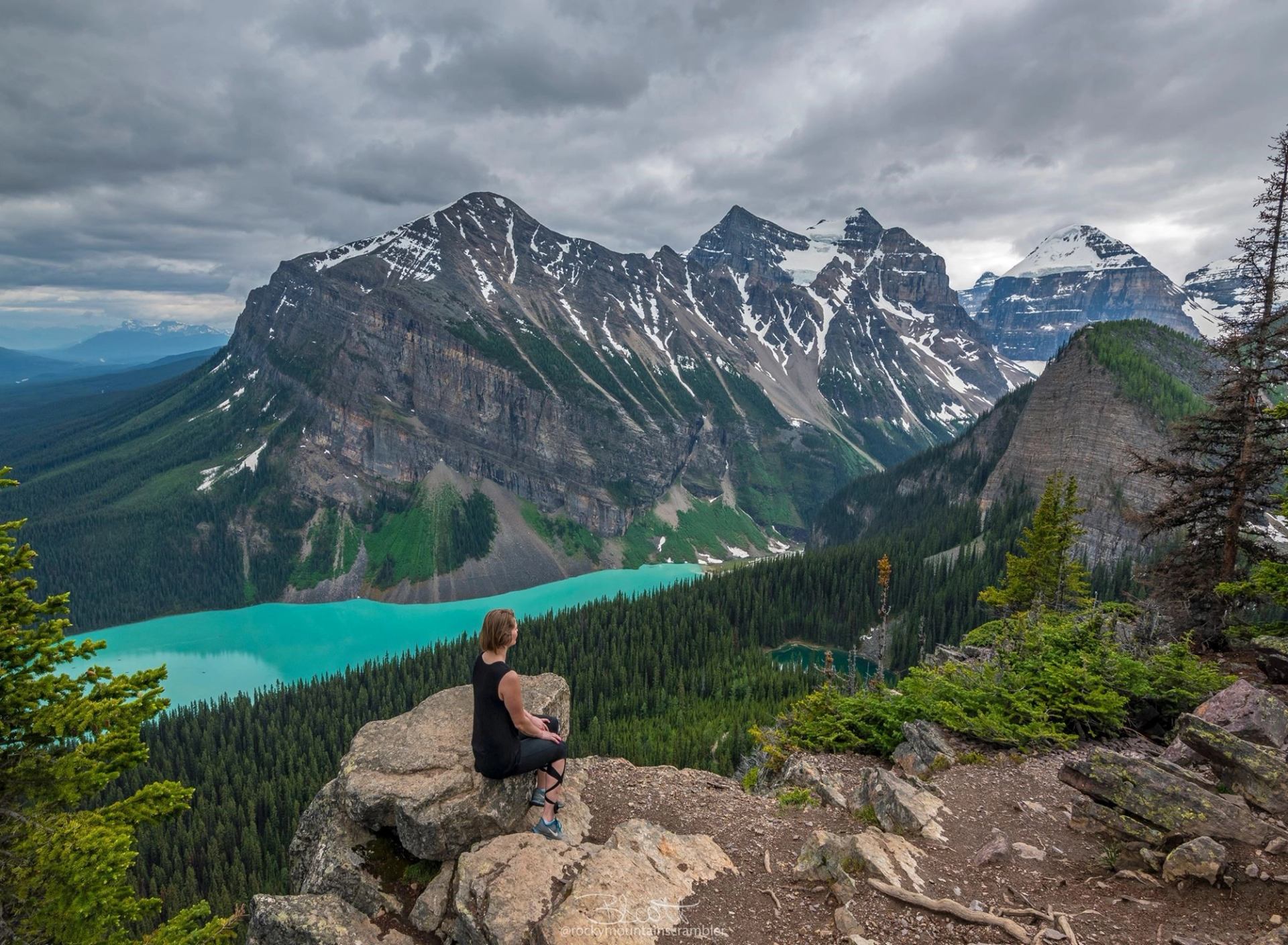 Hiker sitting on rocky cliff overlooking turquoise lake, dense forest and snow-capped mountains.