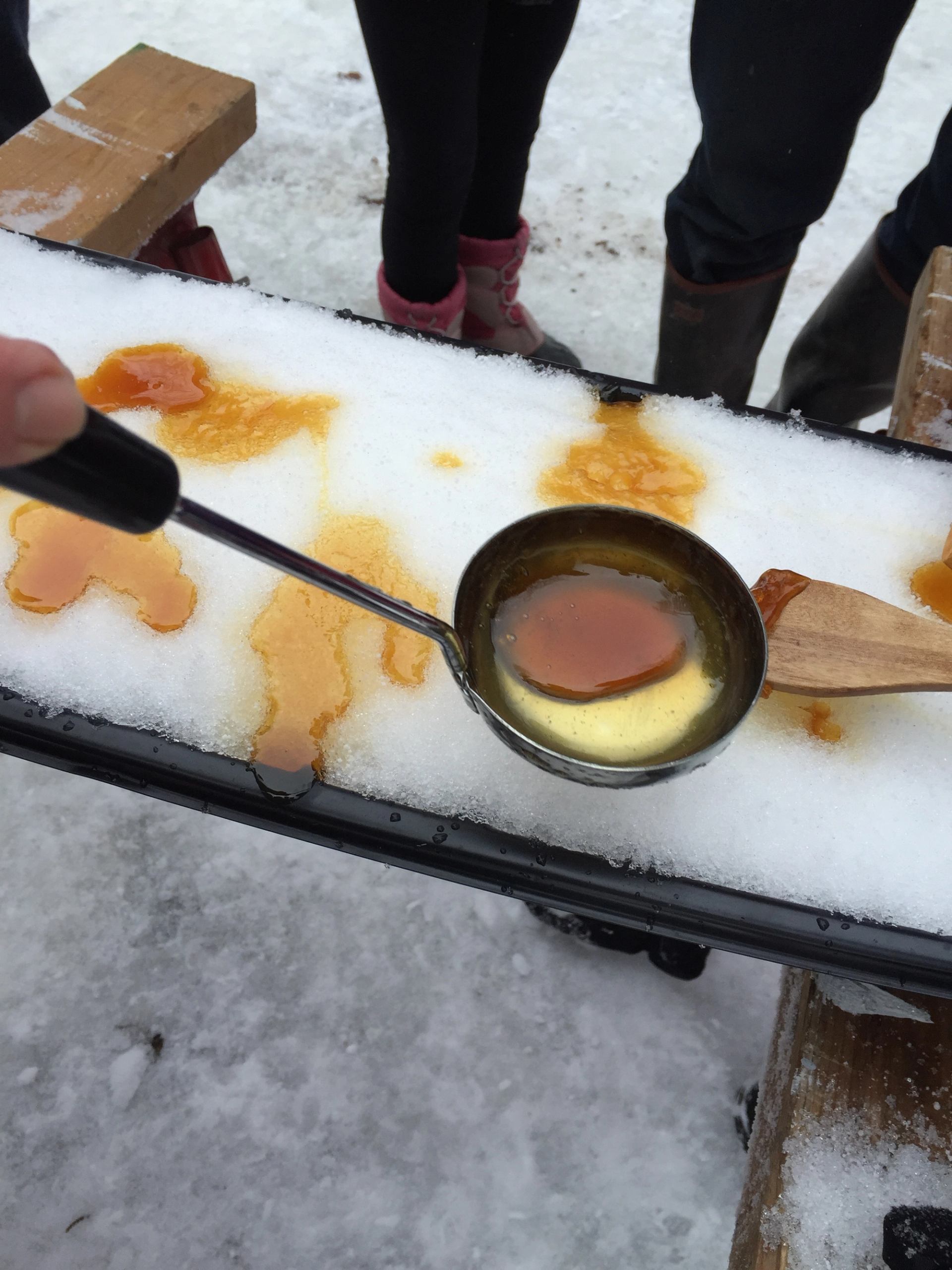 A person is pouring hot maple syrup from a ladle onto a bed of snow to make maple taffy, with boots and legs of other people visible in the background.