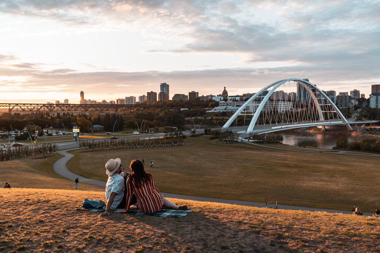 Two people sit on a grassy hill, overlooking a city skyline with an arched bridge over a river at sunset.