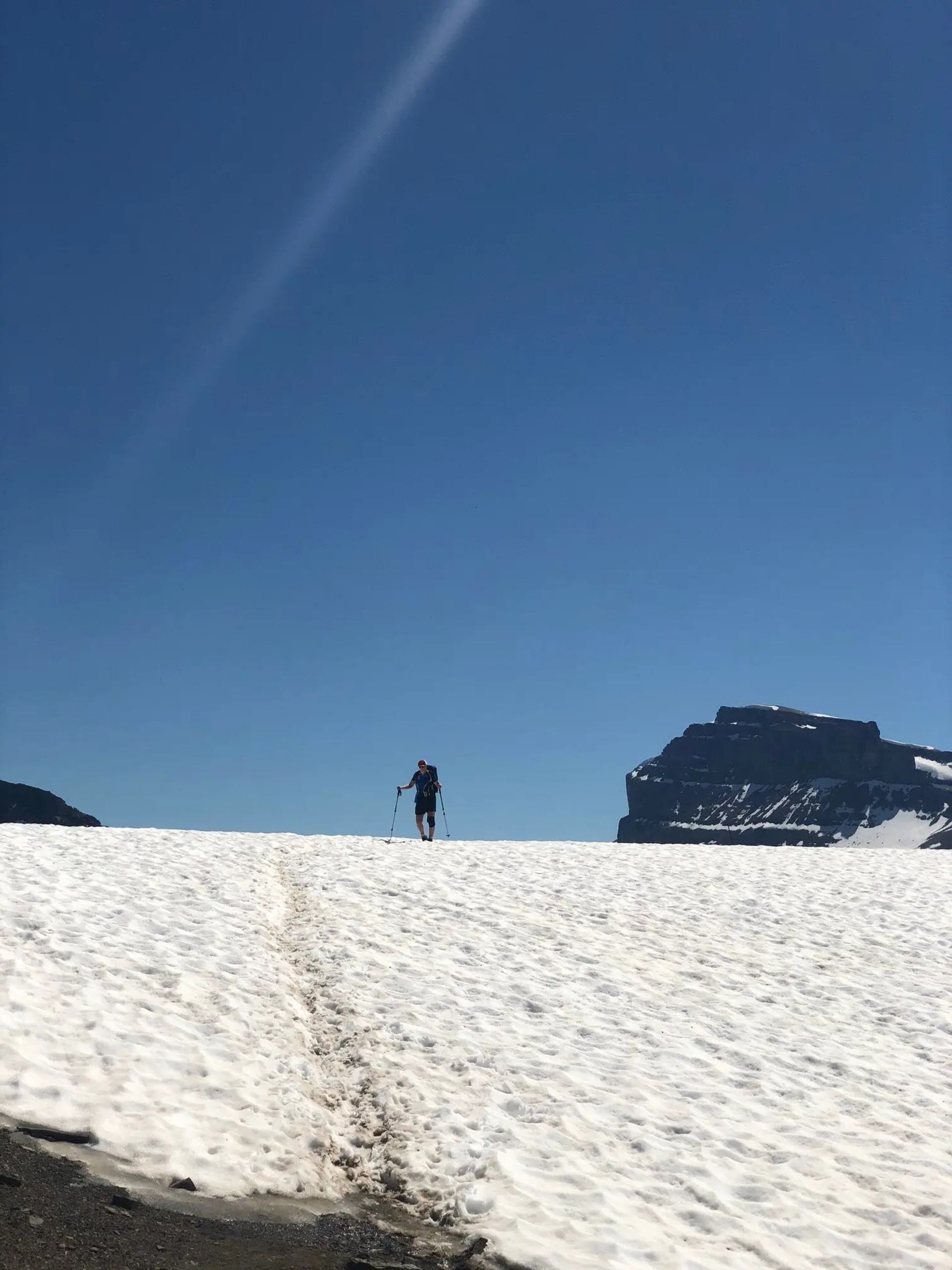 Hiker with trekking poles crosses a snowfield beneath a blue sky and rocky ridge.