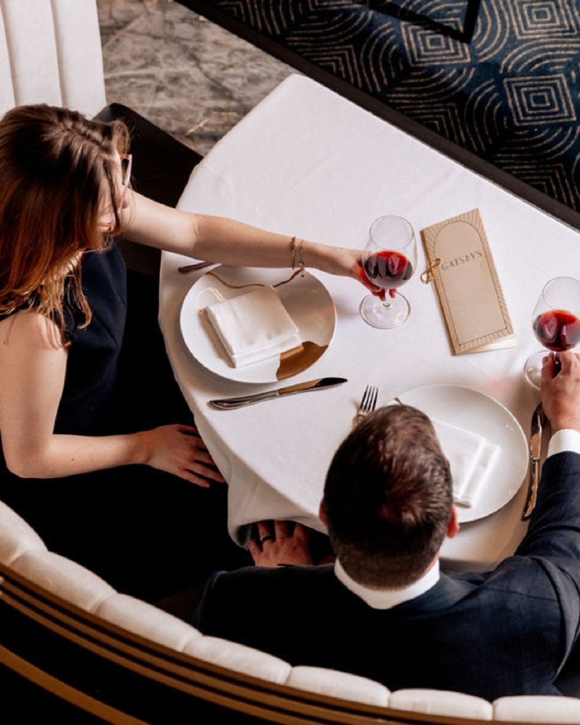 Two people at a restaurant table clink red wine glasses beside menus and plates.