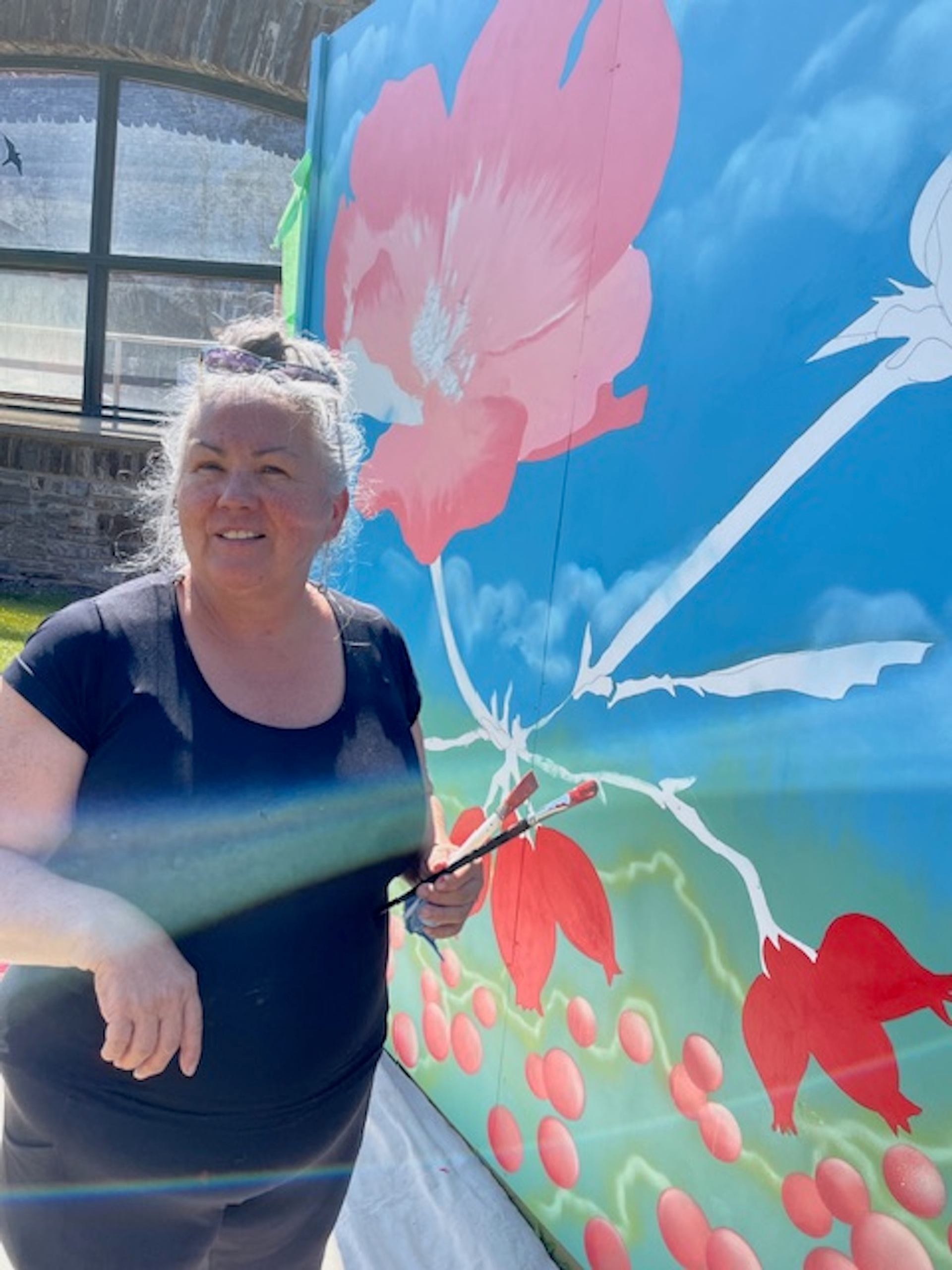 Artist painting a vibrant mural with large pink flowers and green stems on a bright blue background.
