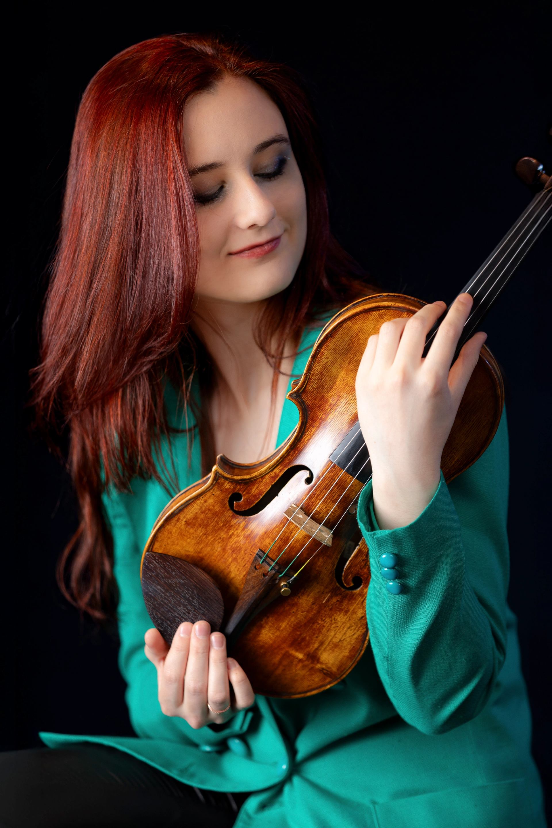 Red-haired person in a teal jacket holds a violin against a black background.