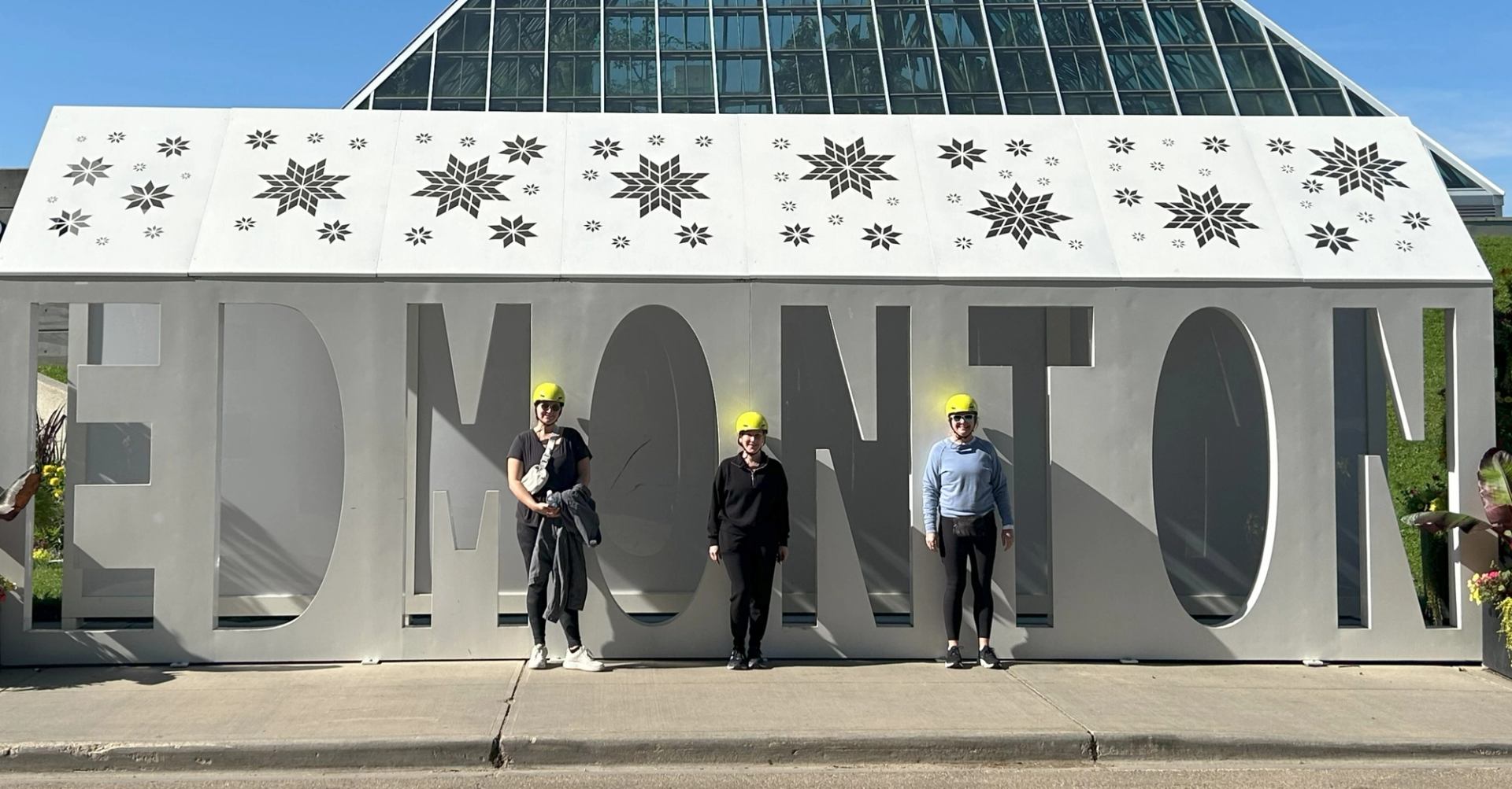 Four people pose in and around large “EDMONTON” sign with snowflake decor and blue sky.