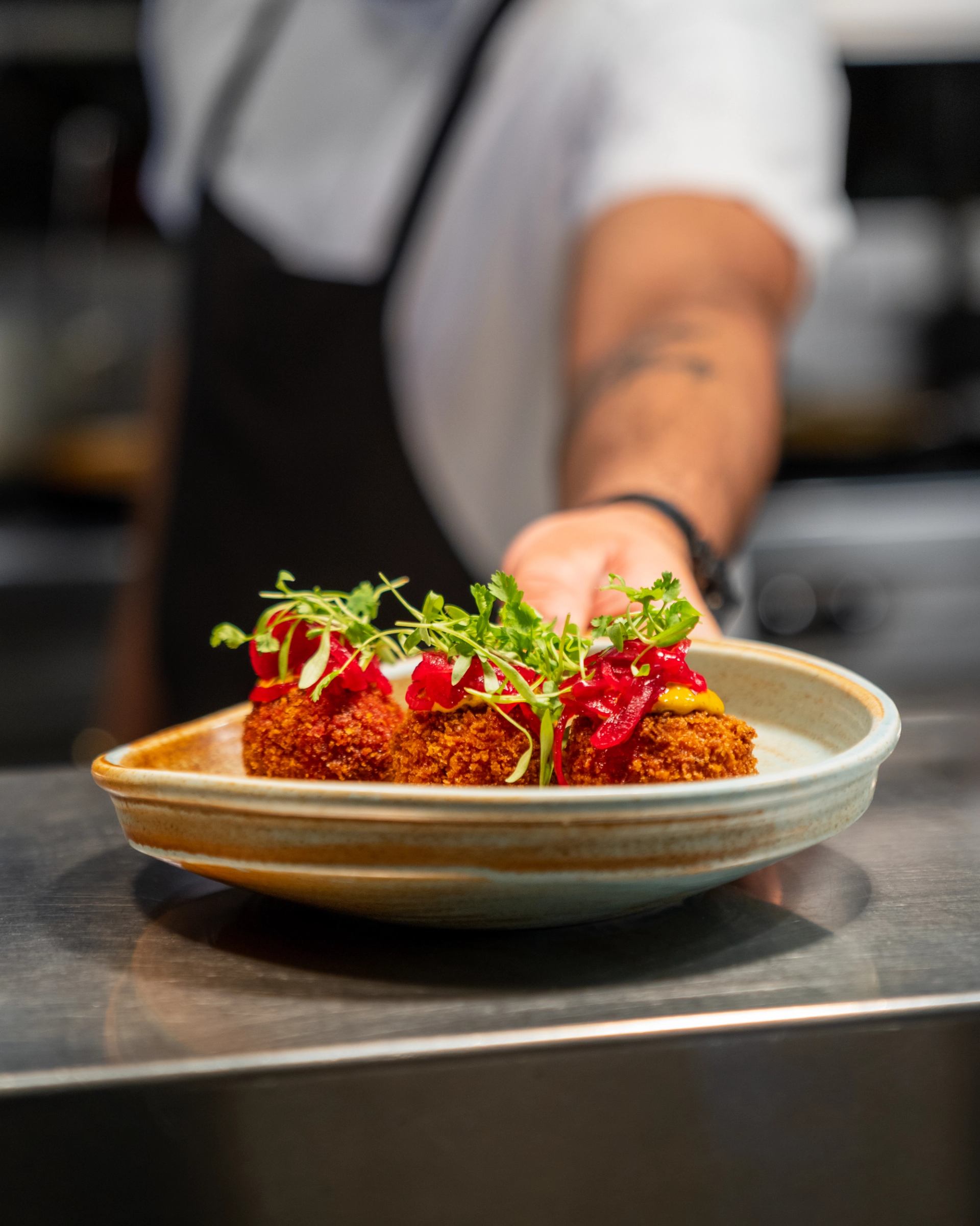 Chef presents a plate of breaded croquettes topped with greens and red garnish.