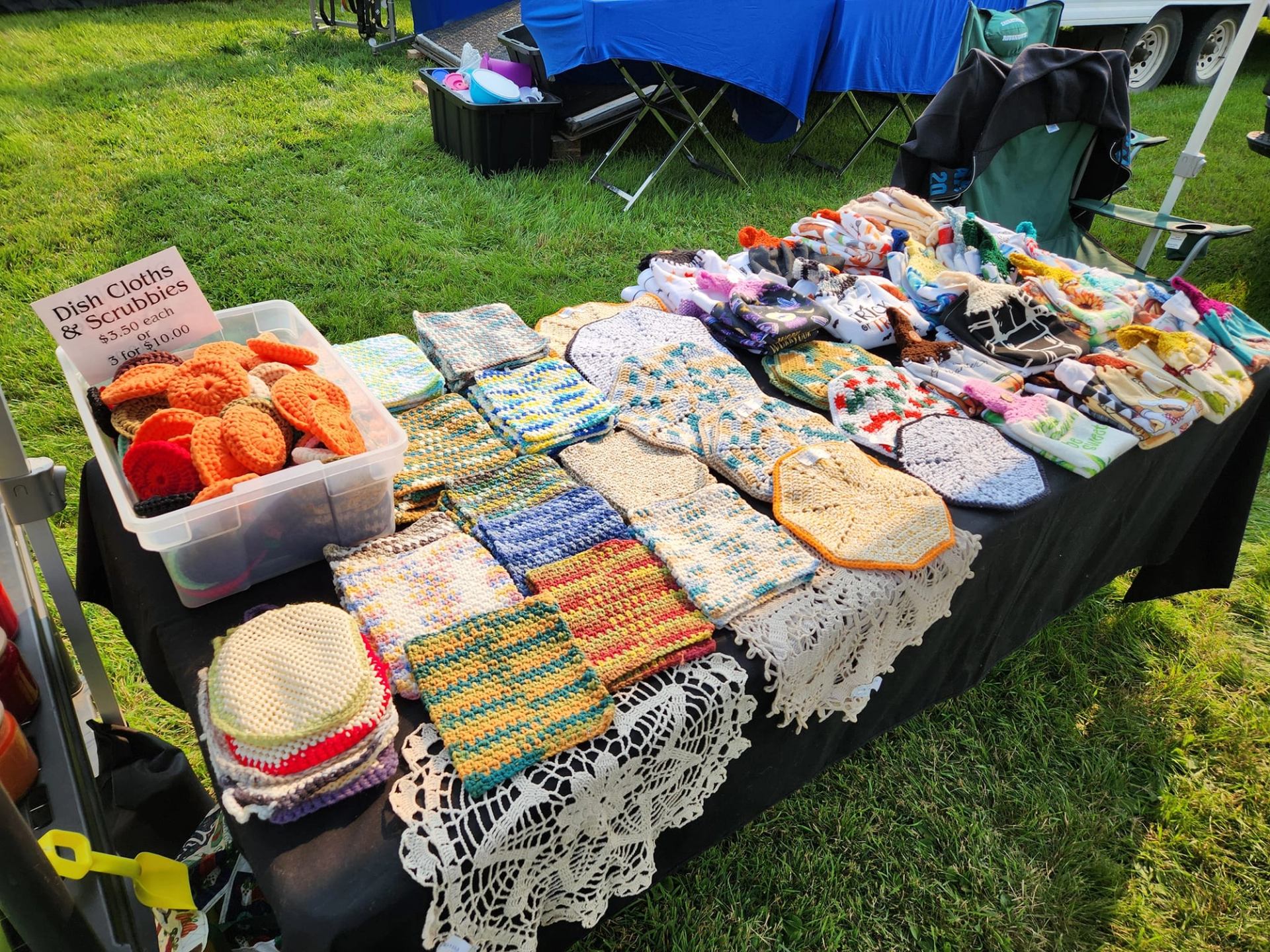 Handmade crocheted dishcloths, scrubbers, and crafted goods are displayed on a vendor table outdoors.