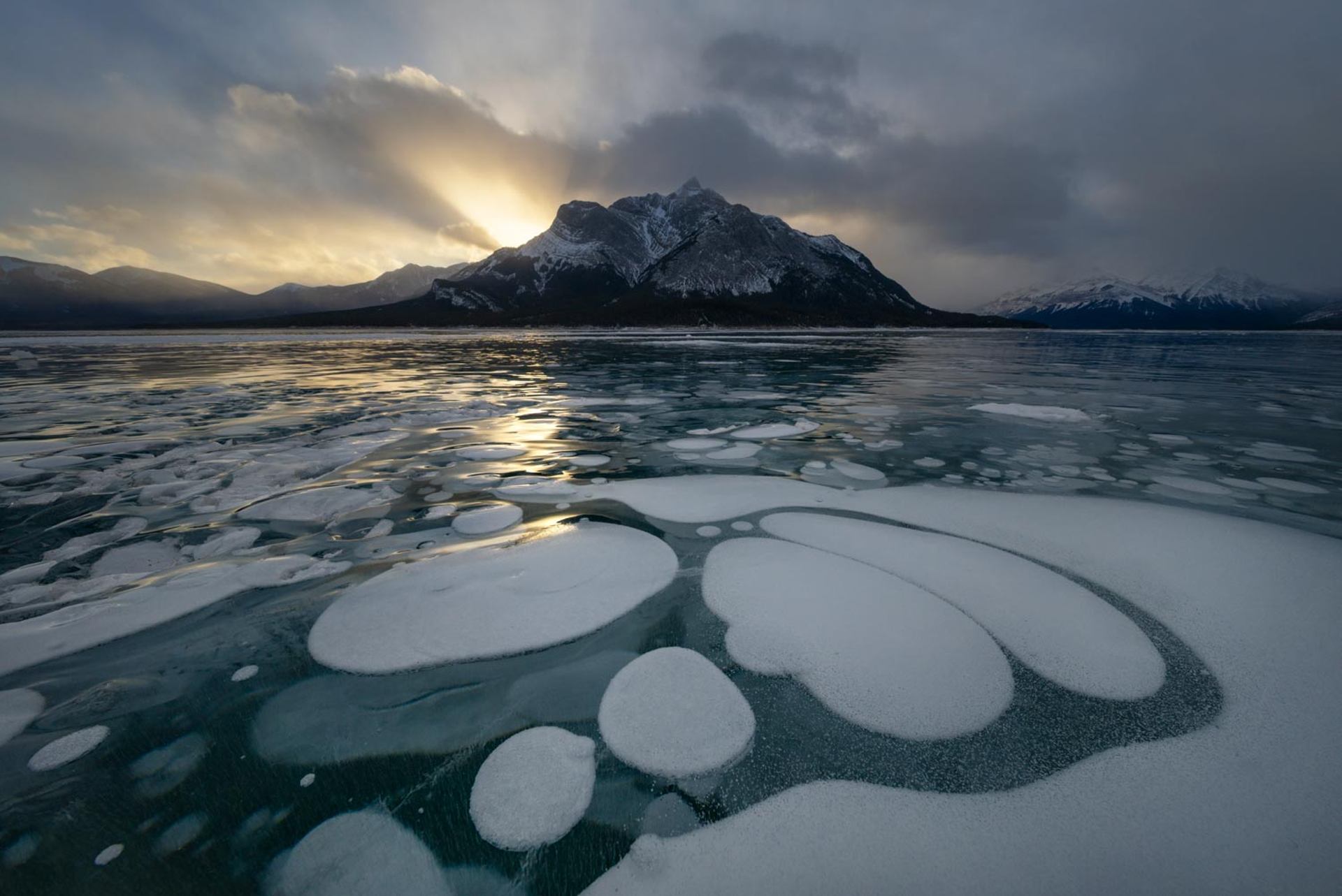 A frozen lake with trapped white bubbles in the ice, a dark mountain, and sun rays breaking through dramatic clouds.