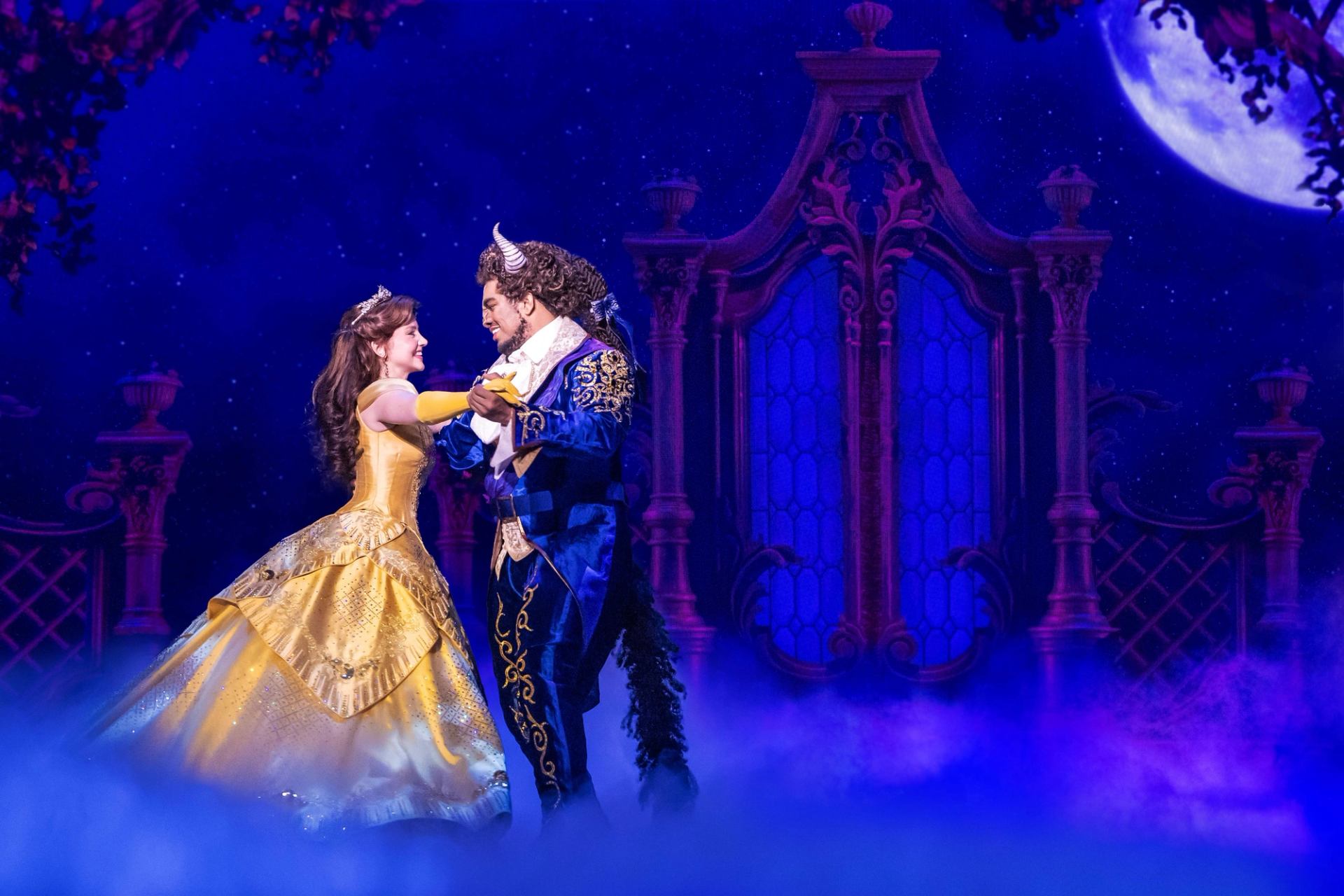 Stage actors as Belle and Beast dance in ornate ballroom set with blue lighting.