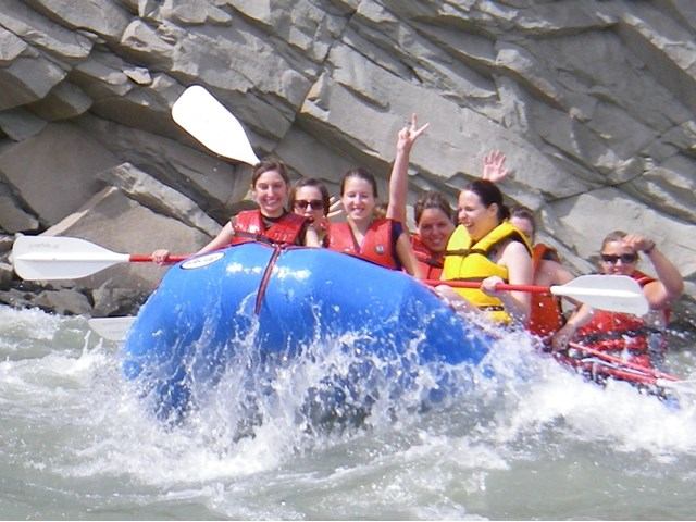 Raft hitting whitewater rapids as paddlers lean in against rushing waves near rocky cliffs.