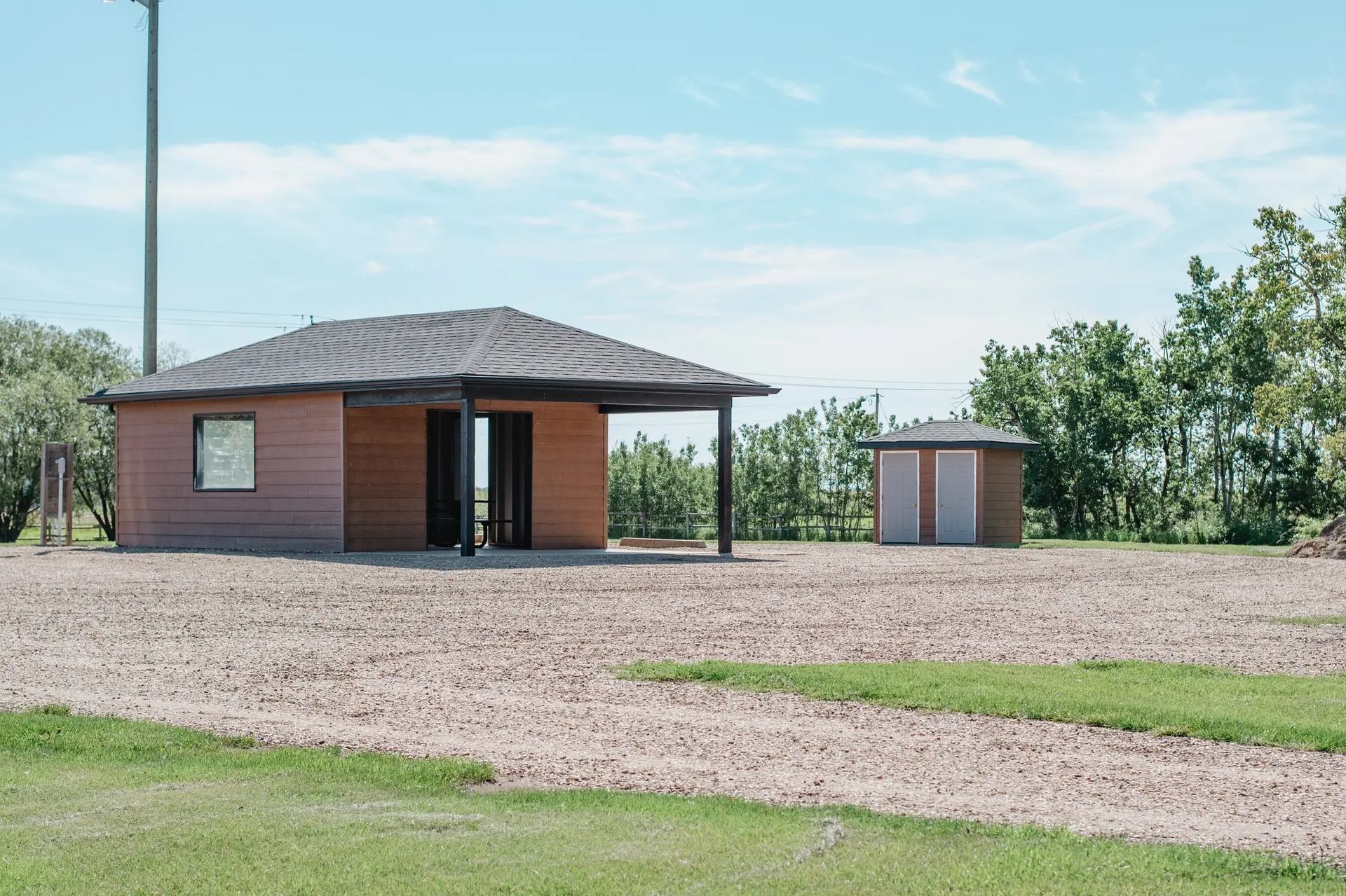 Small brown building with covered entrance beside shed on gravel lot and grass.