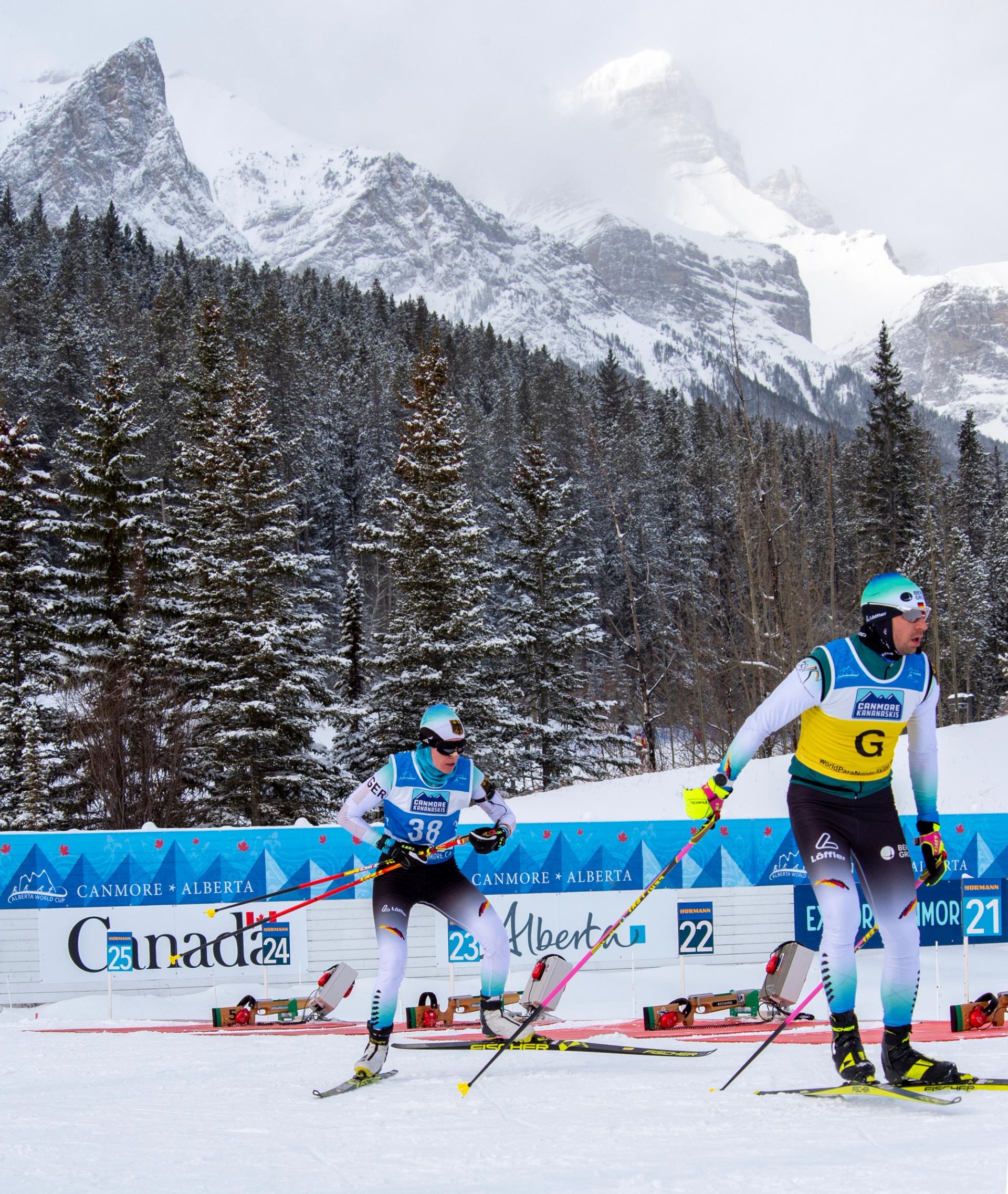Two para biathletes skiing through snowy mountain terrain.