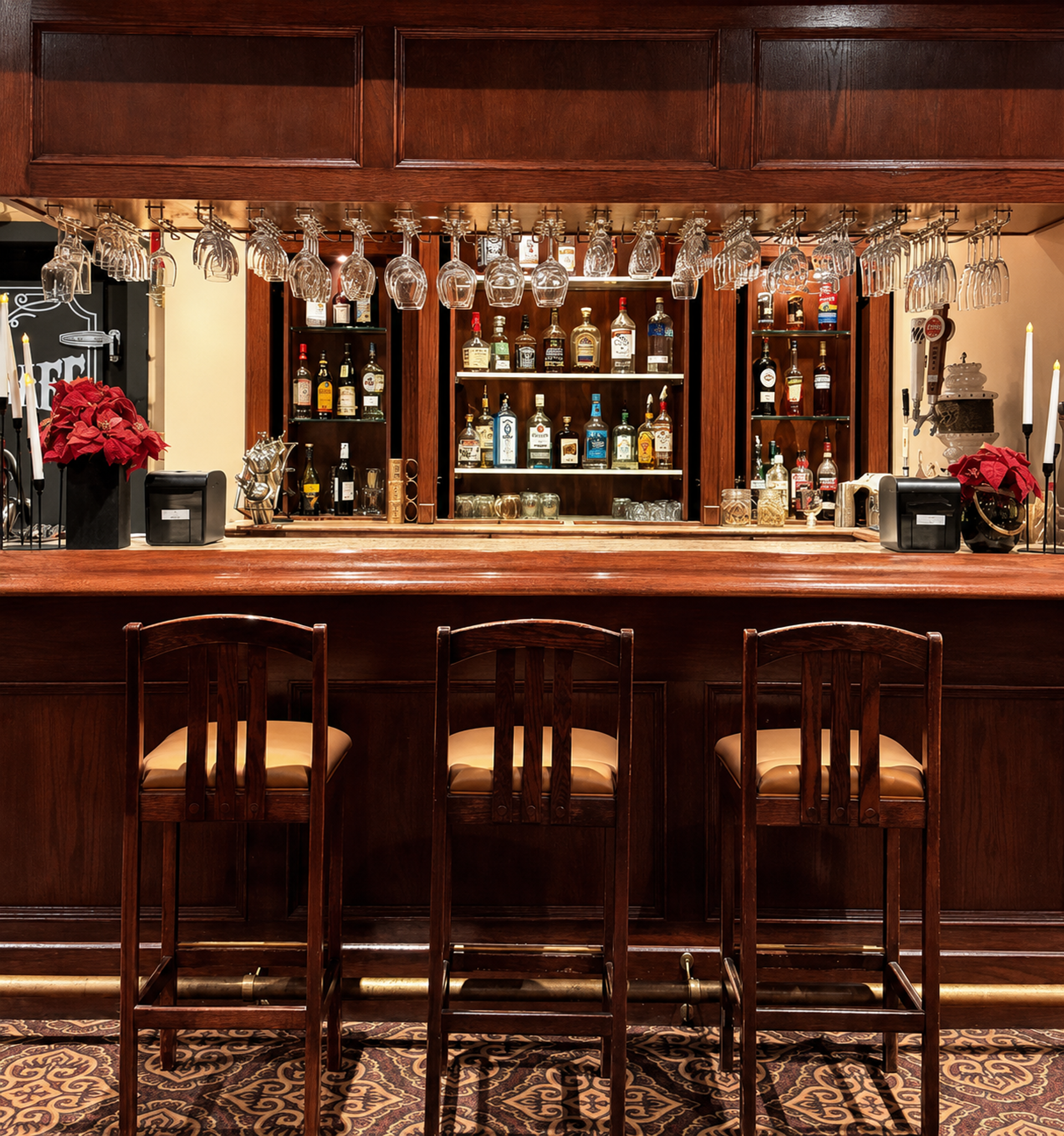 Wooden bar with three stools, hanging wine glasses and liquor bottles on shelves.
