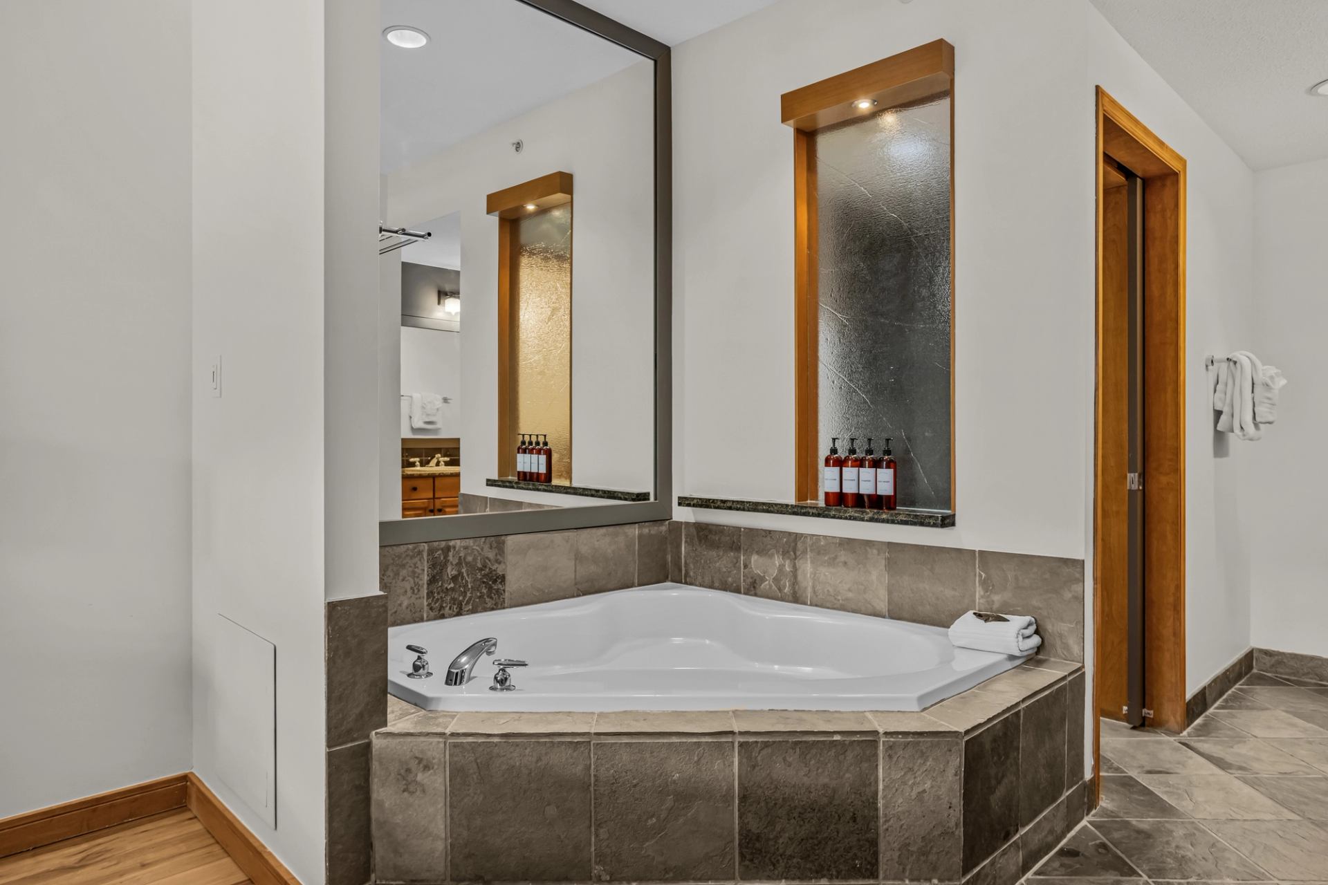 A large corner soaker tub is surrounded by tile accents, mirrors, and built-in shelves with bath amenities.