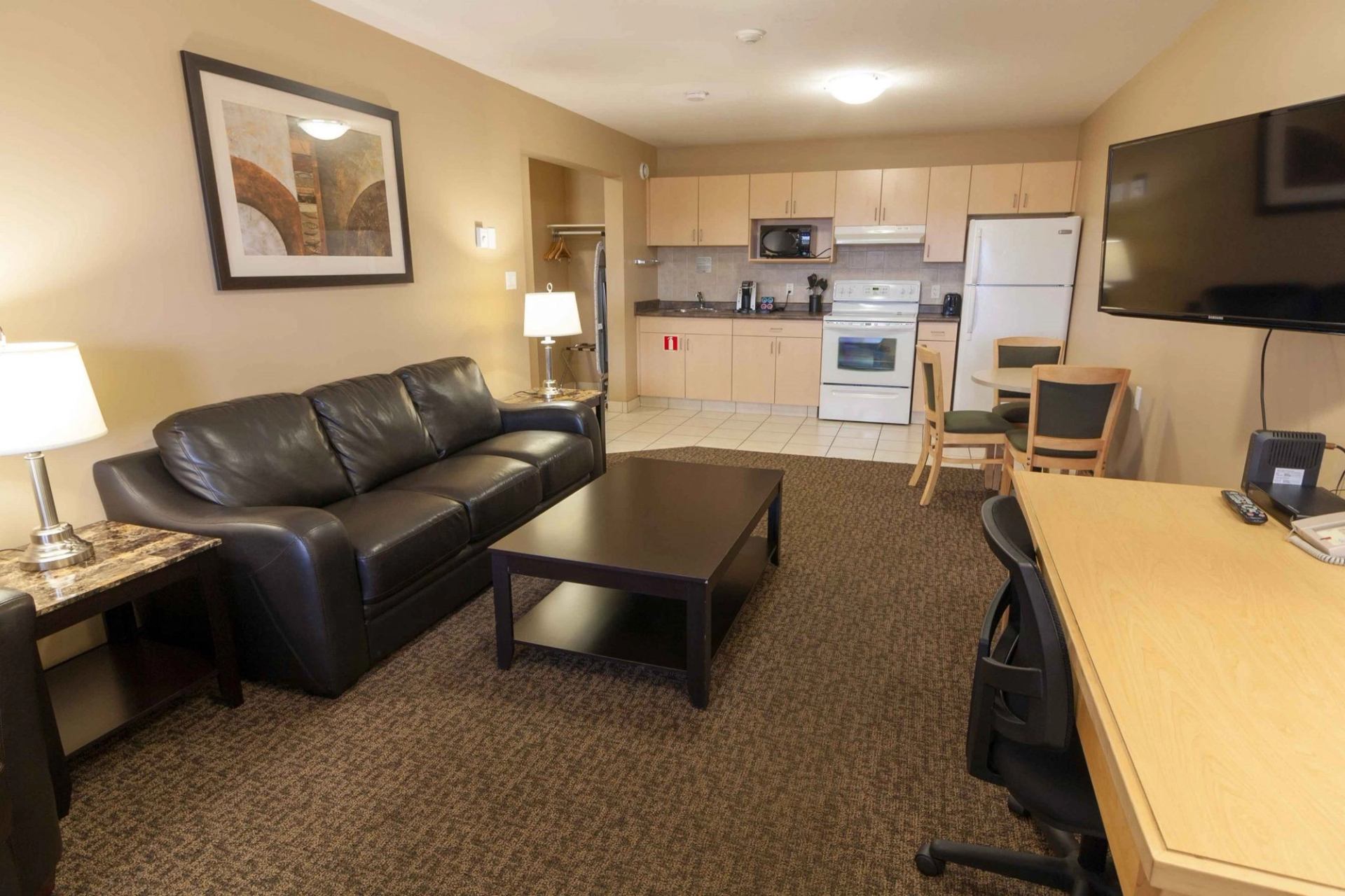 Hotel suite with leather sofa, coffee table, kitchenette, dining table, desk and wall-mounted TV.
