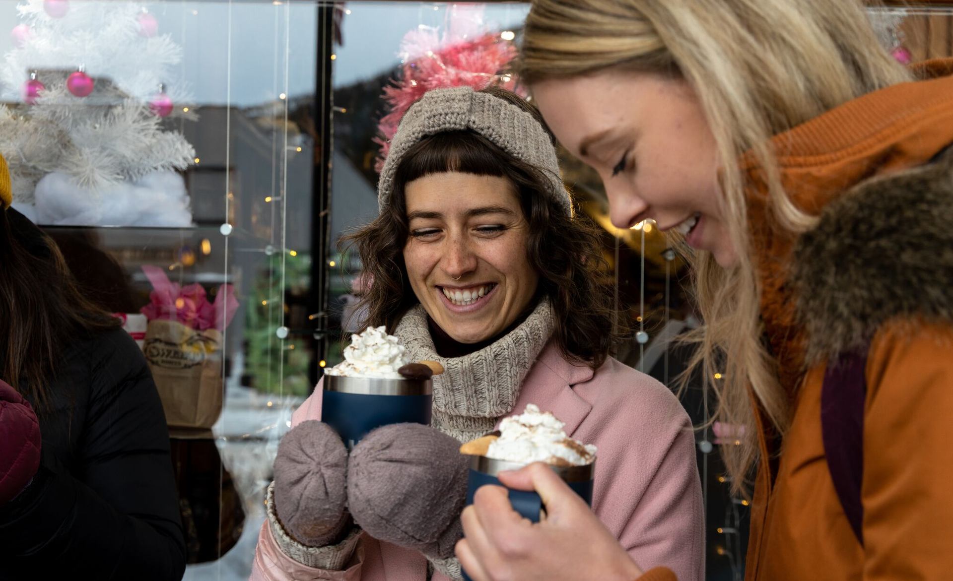 Two people enjoying whipped cream-topped hot chocolate outdoors in winter attire.