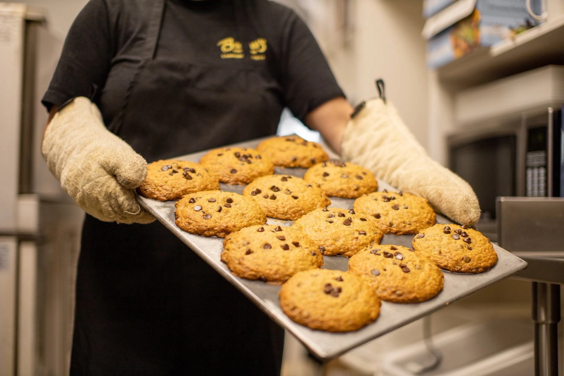 Person holding a tray of fresh chocolate chip cookies while wearing oven mitts in a kitchen.