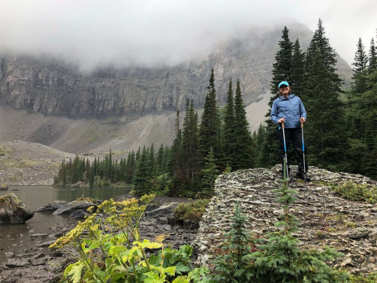 Hiker with trekking poles stands on rocky ledge beside alpine lake and conifer forest.