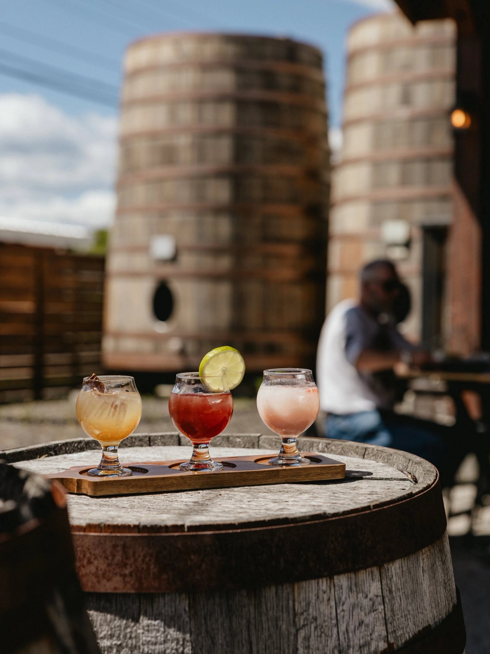 Three handcrafted cocktails sit on a wooden tasting flight atop a barrel, with large aging casks beh