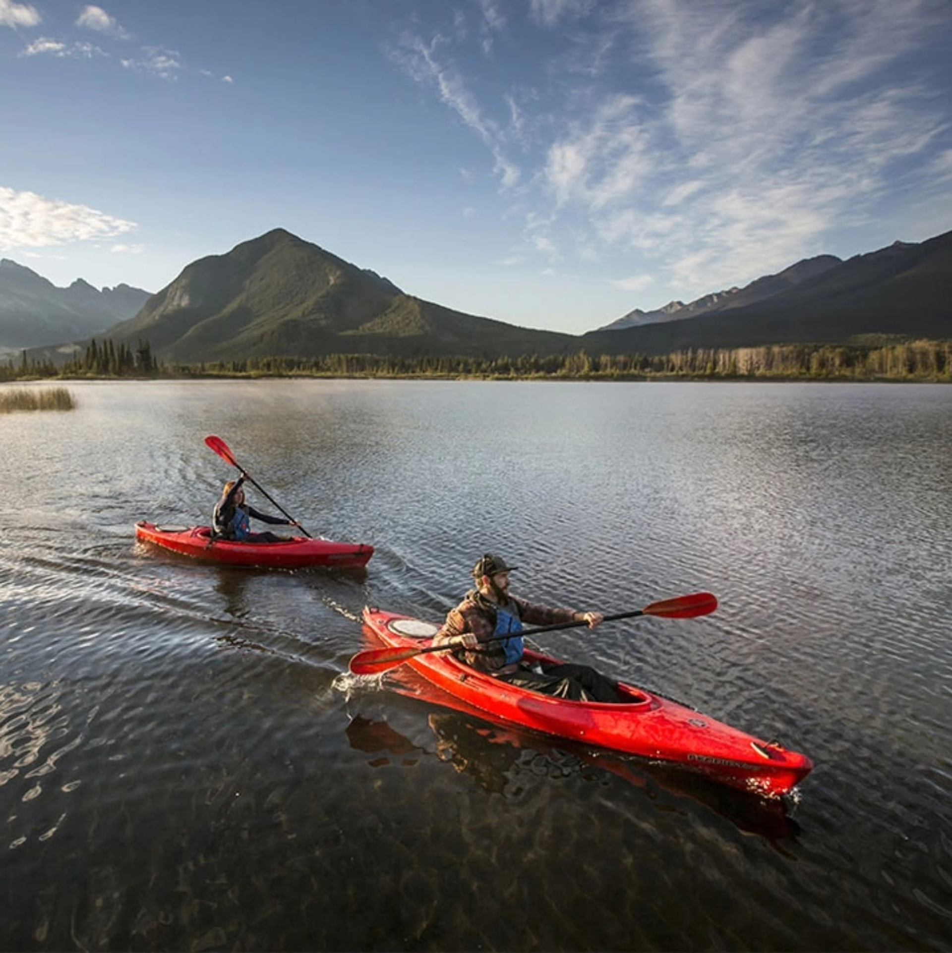 Two people paddle red kayaks across a calm lake with forested mountains behind.
