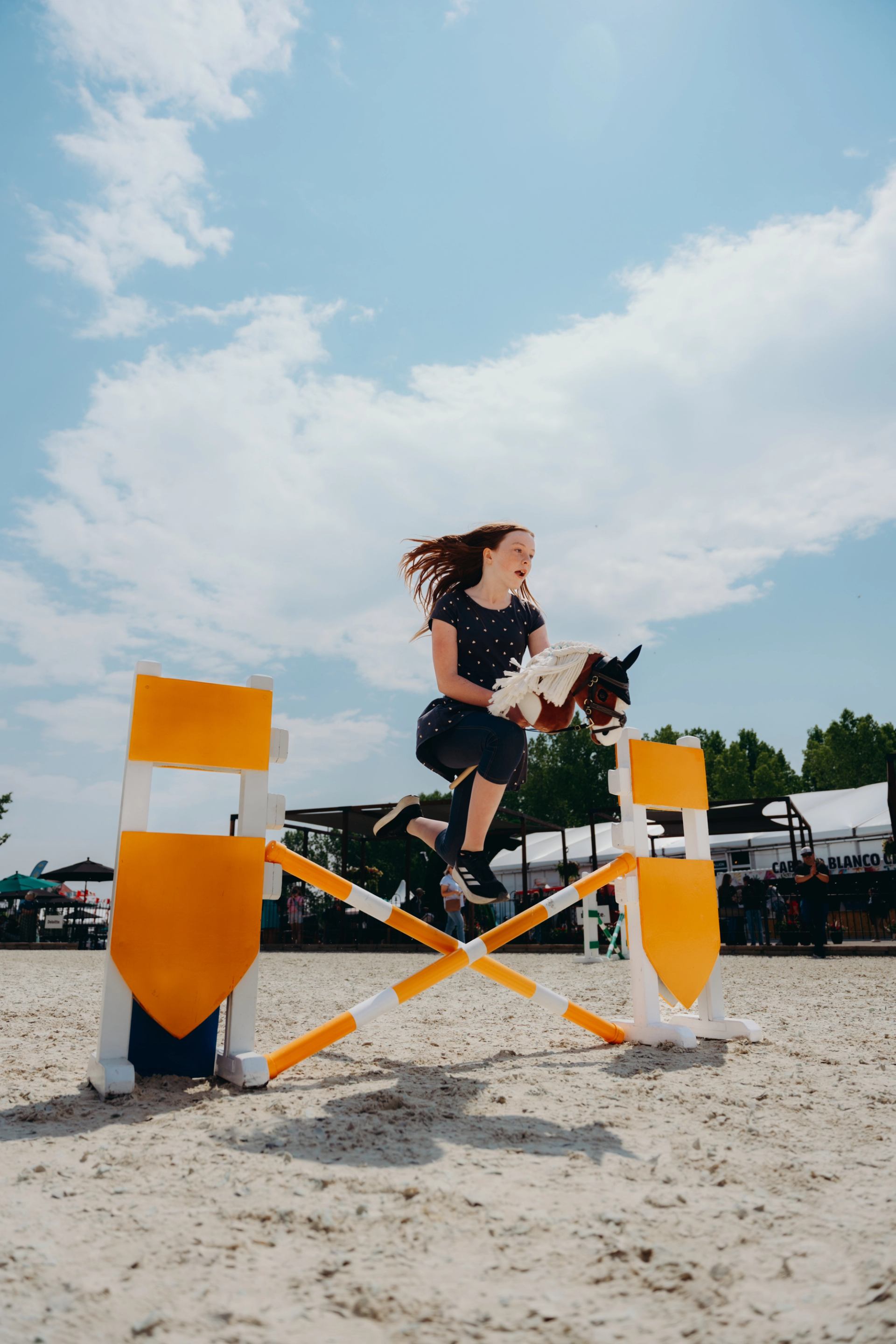 Child jumps over yellow practice hurdle while holding toy horse at outdoor arena.