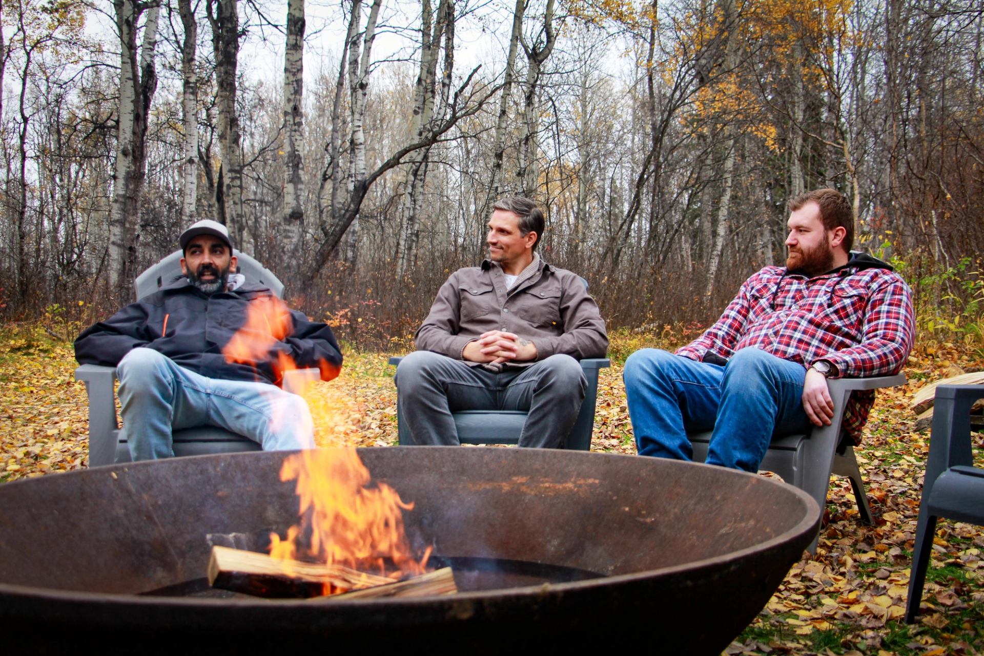 Three people relaxing around a firepit in a forest clearing with autumn leaves.
