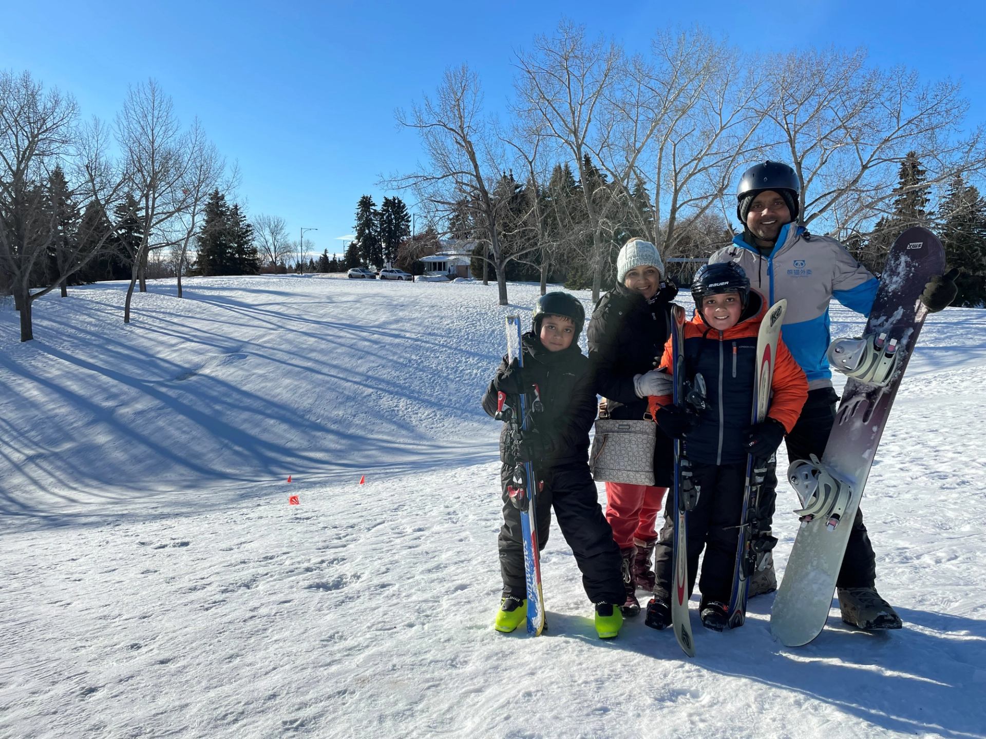 Snowboarders in helmets holding snowboards on a snowy outdoor ski slope with trees and cars.