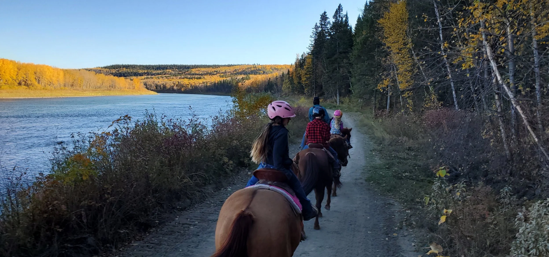 Horseback riders follow a forest trail beside a river and yellow-leaved hills.
