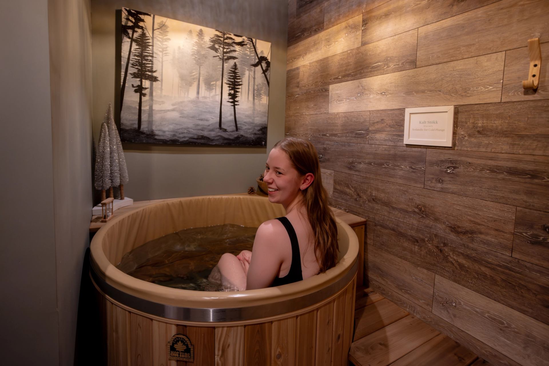 A smiling woman sits inside the wooden cold plunge tub, looking back over her shoulder toward the camera. The space is decorated with a serene forest painting and natural wood elements.