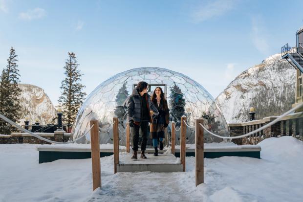 A happy couple dressed in outdoor winter clothing walks out of the geodome and onto the pathway outside at the Fairmont Banff Springs