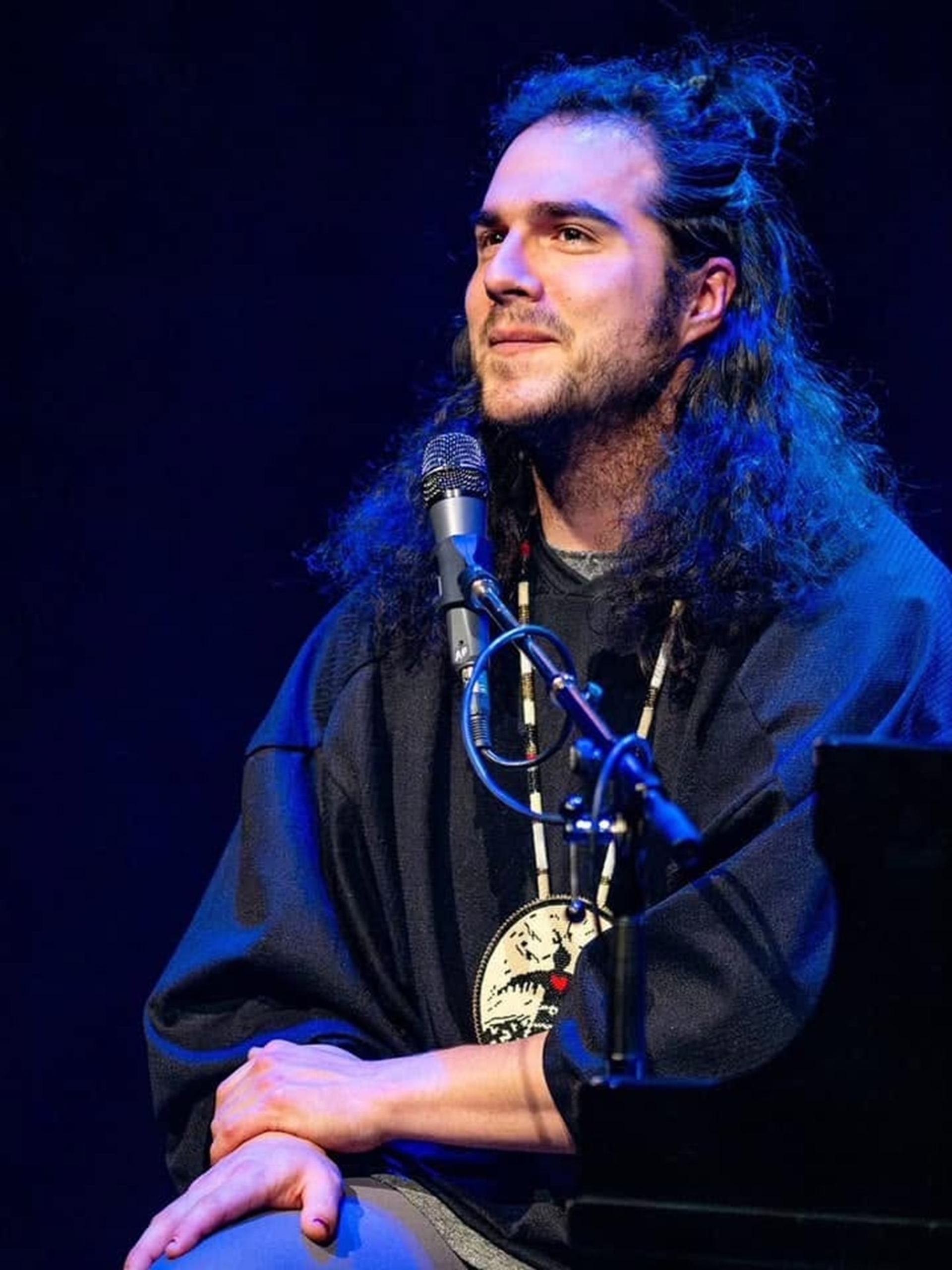 Long-haired performer sits at a microphone under blue stage lighting.