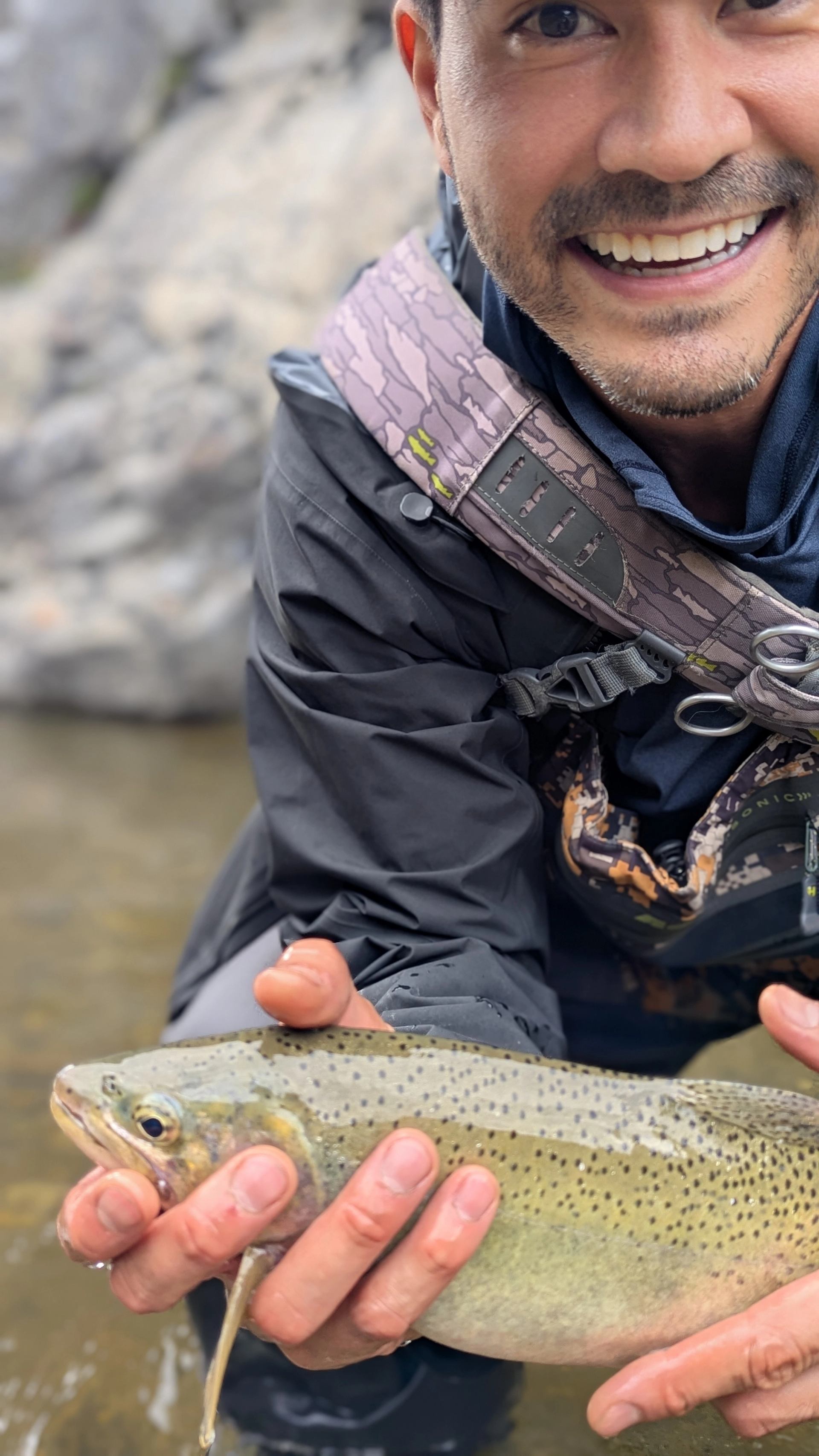 A smiling man in fishing gear holds a trout in the water.