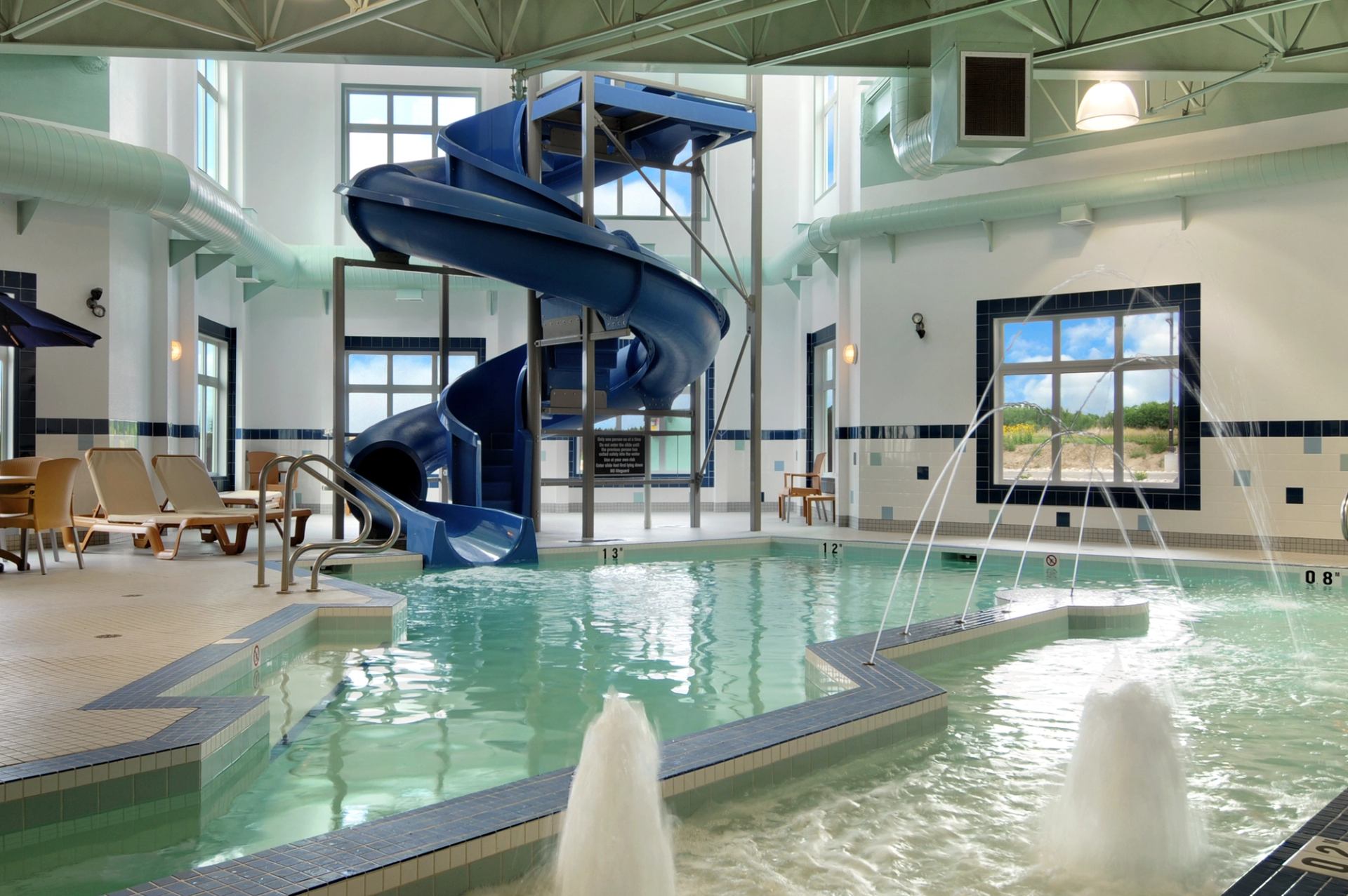 Indoor hotel pool with blue waterslide, water fountains, lounge chairs and large windows.