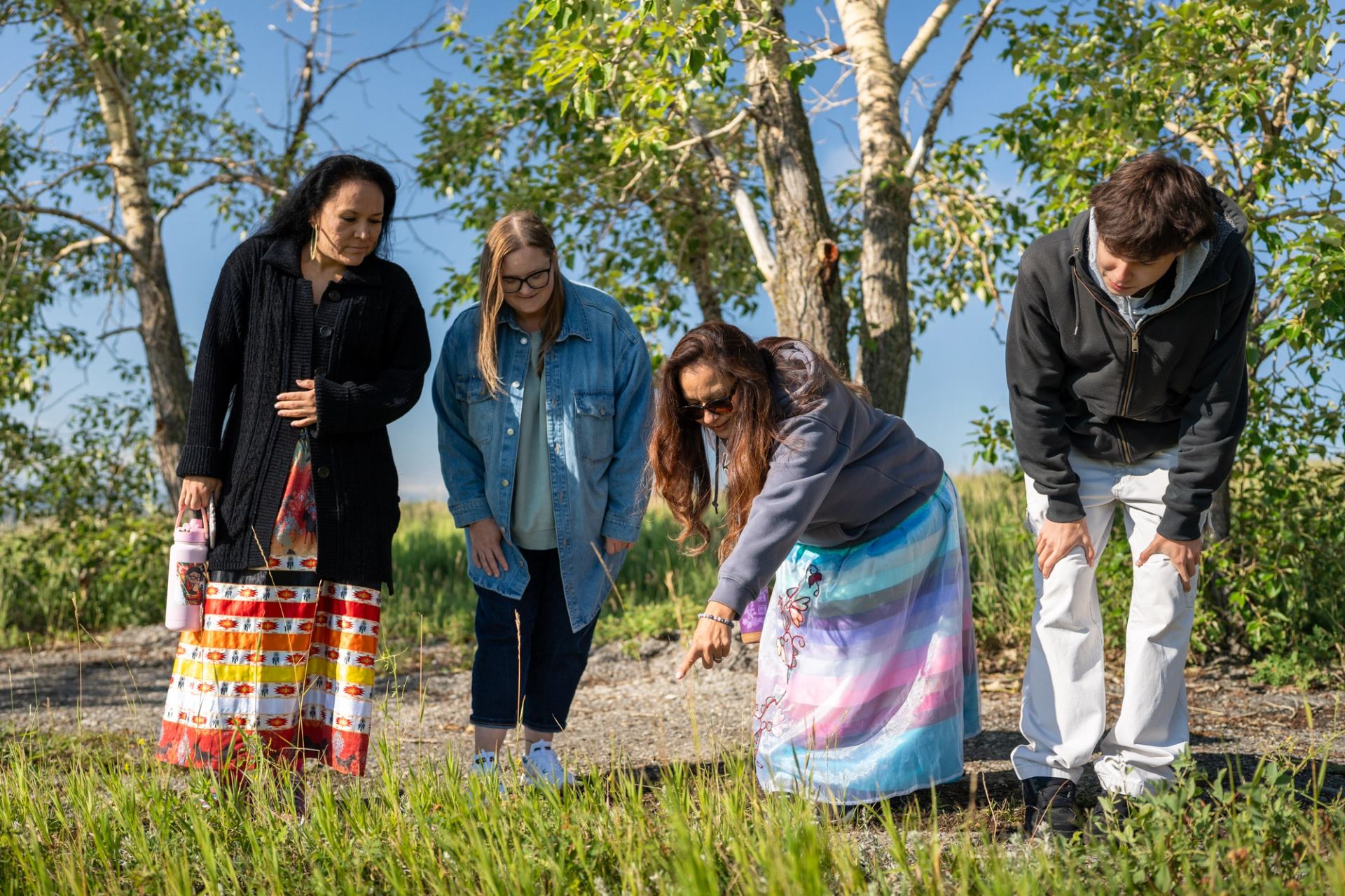 A small group stands outdoors, looking down and connecting with the land among trees.