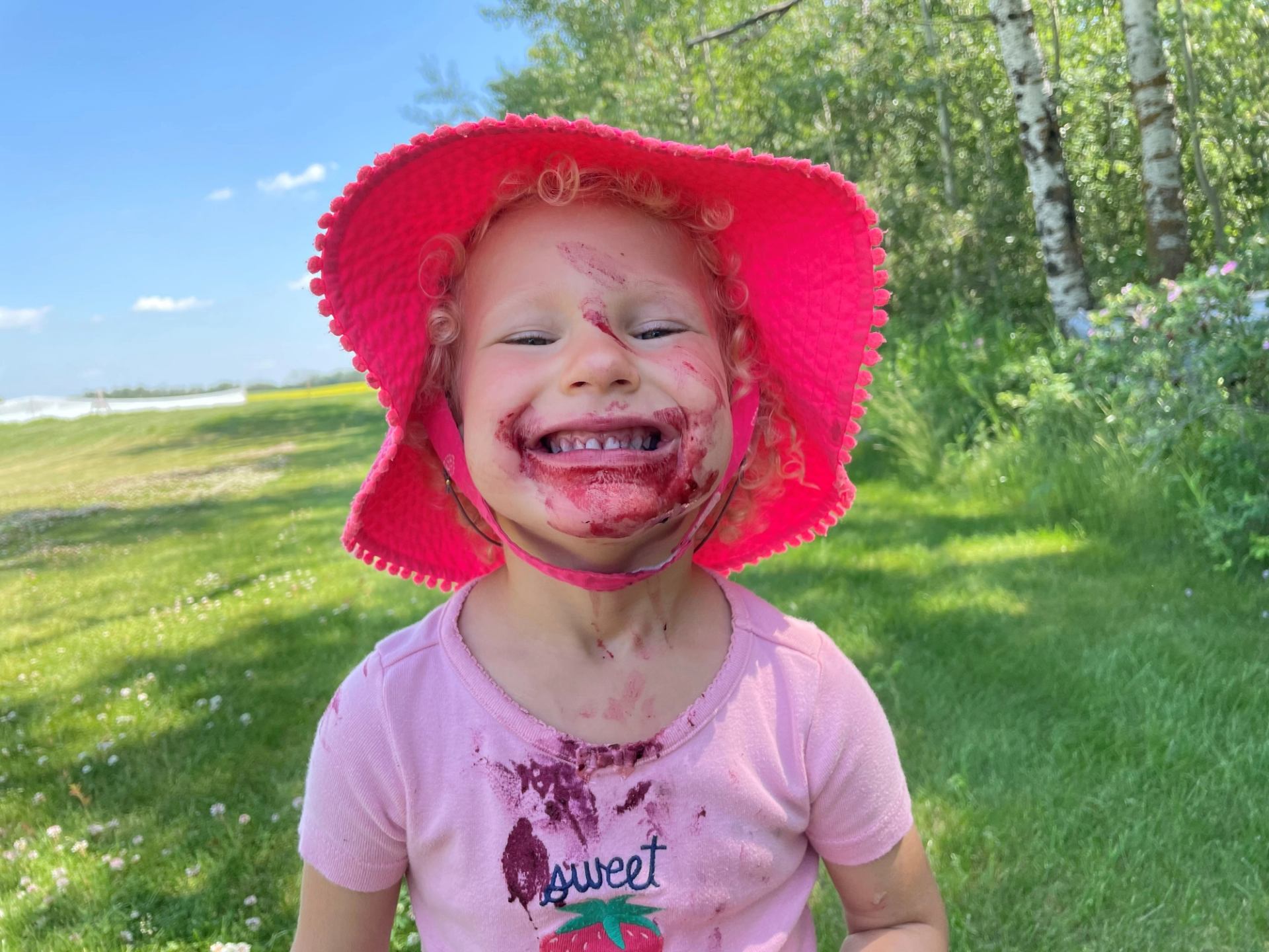 Child with berry-stained face wearing bright pink sun hat in grassy park.