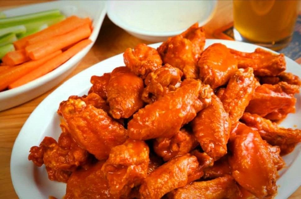 Plate of sauced chicken wings with celery, carrot sticks, dipping sauce and beer.
