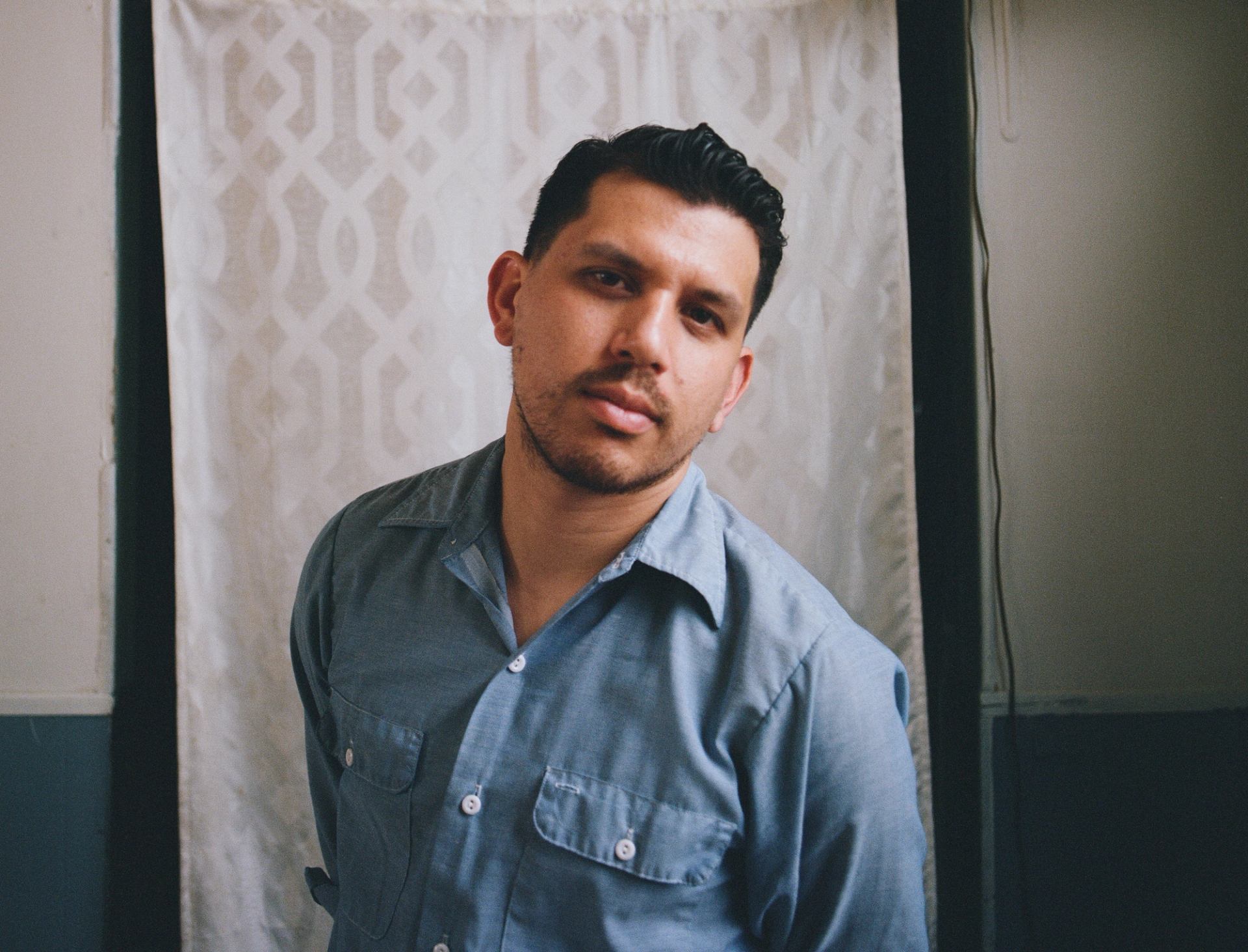 A man in a blue shirt with dark hair and a light beard looks at the camera against a patterned white curtain.