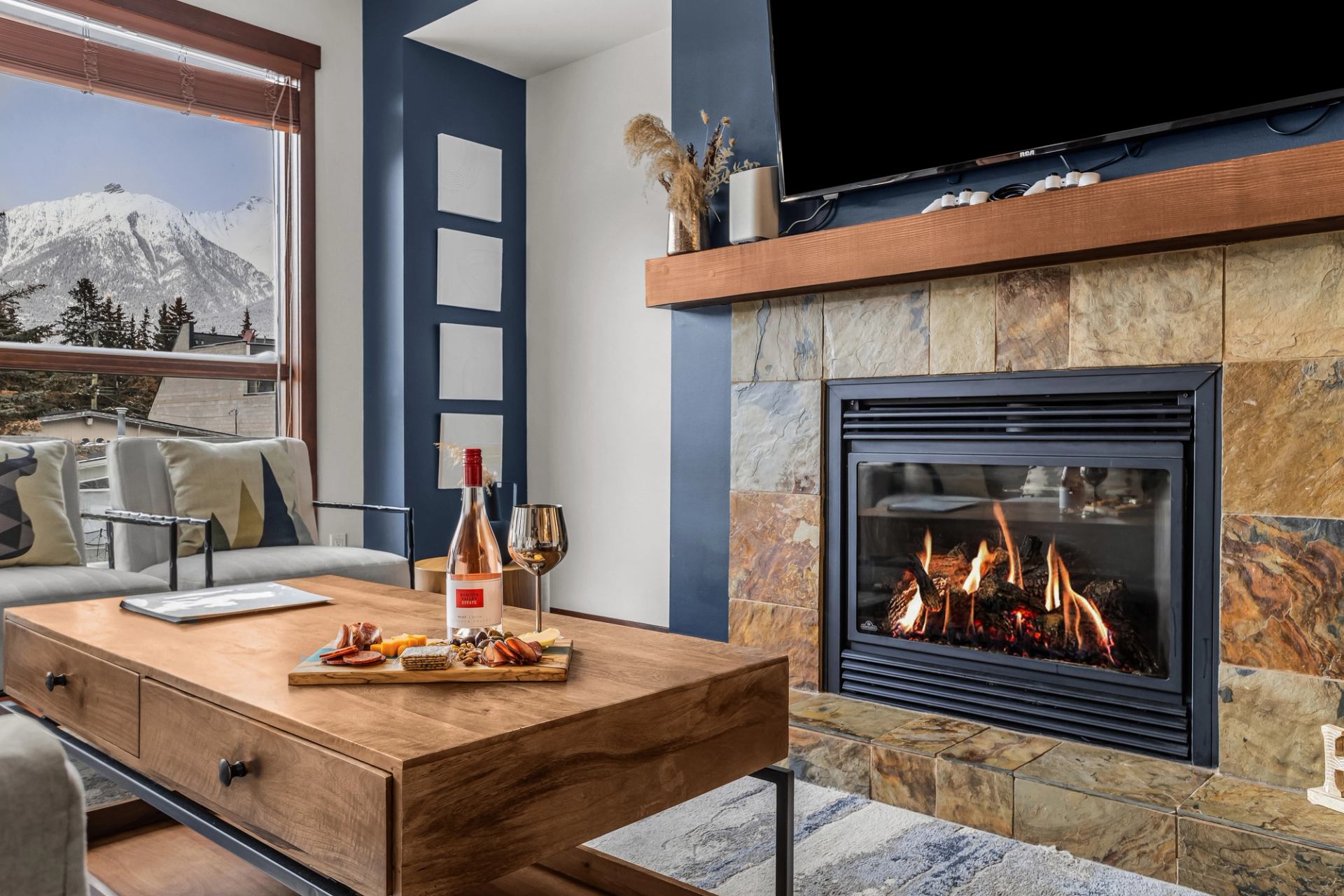 Living room with lit stone fireplace, wine and snacks on wooden coffee table.