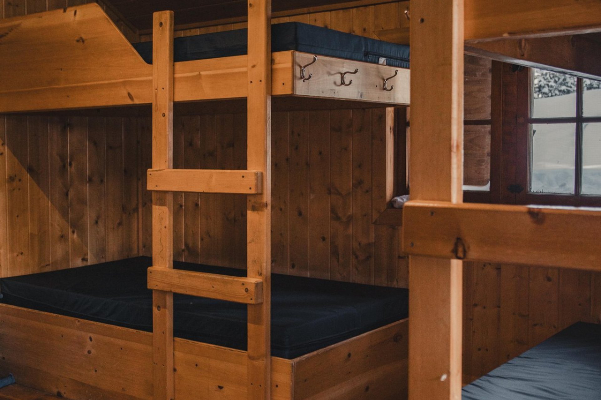 Dorm room with wooden bunk beds, navy blue mattresses, and wall hooks for gear.