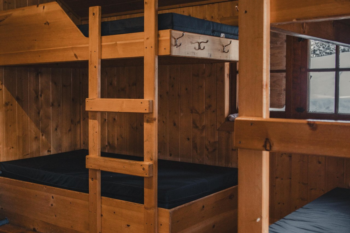 Dorm room with wooden bunk beds, navy blue mattresses, and wall hooks for gear.