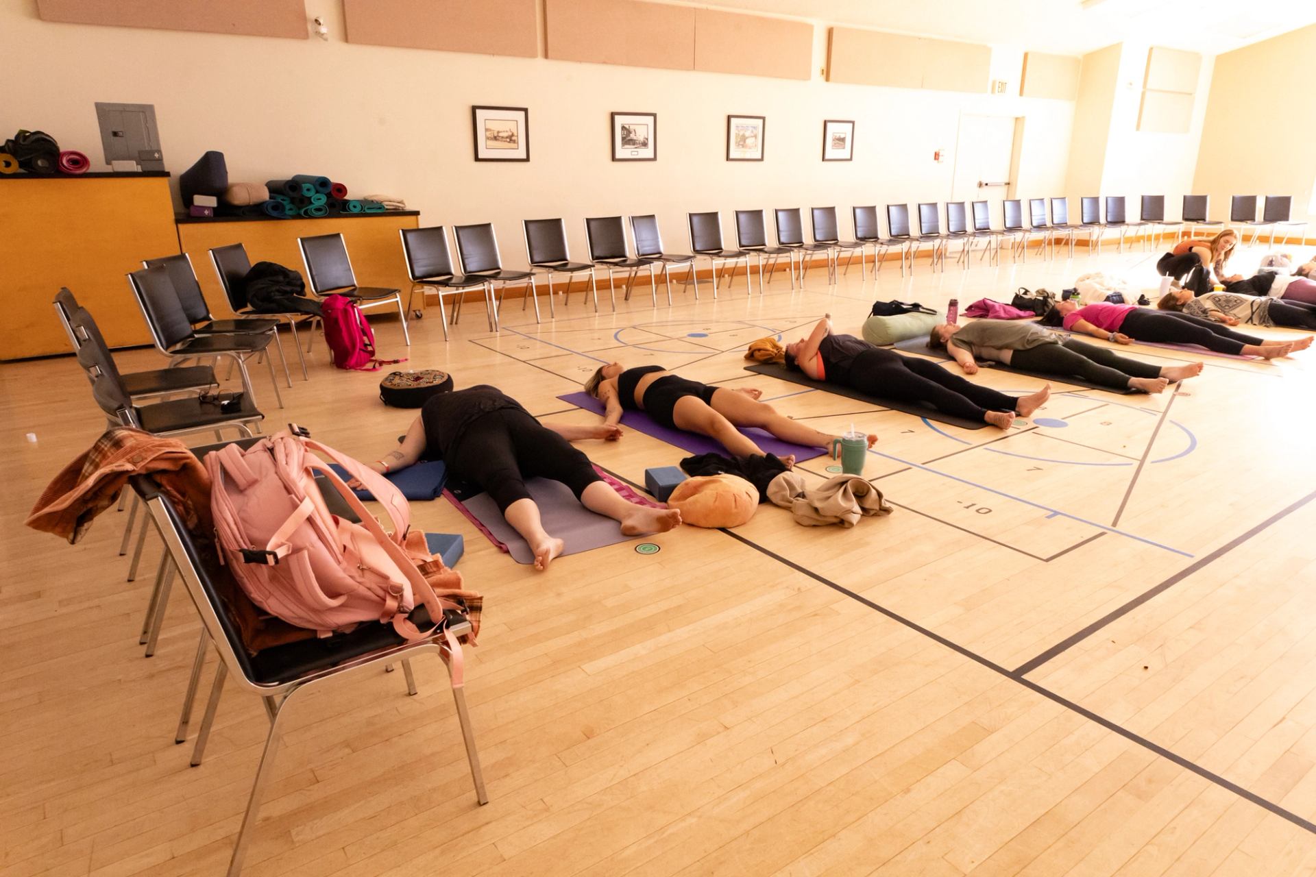 People lie on yoga mats in a gym, surrounded by chairs, bags and props.