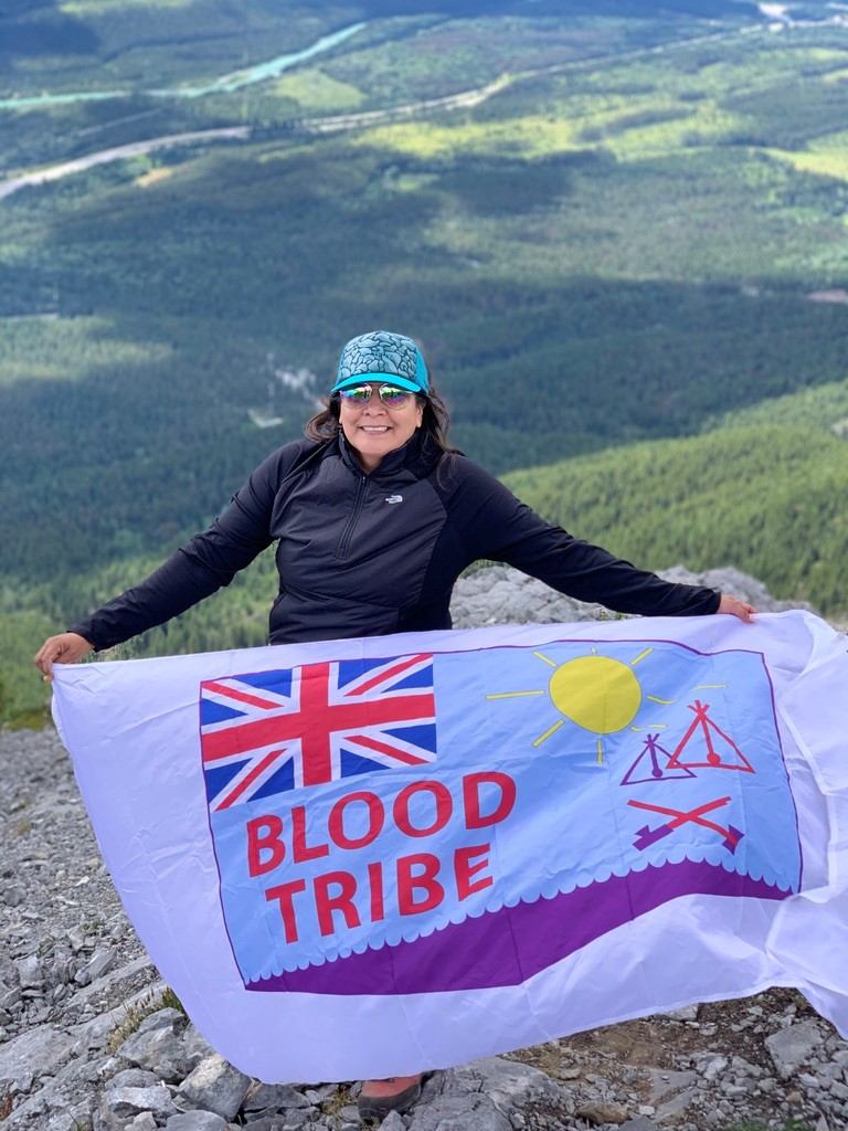 Person holding a Blood Tribe flag on a rocky mountain ledge with a valley view below.
