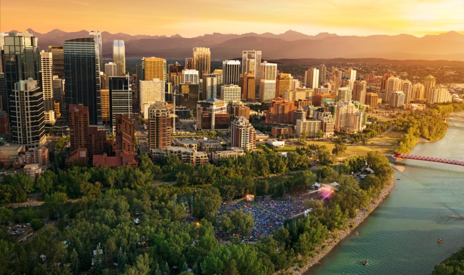 Aerial view of downtown skyline, riverside park, outdoor crowd and distant mountains.