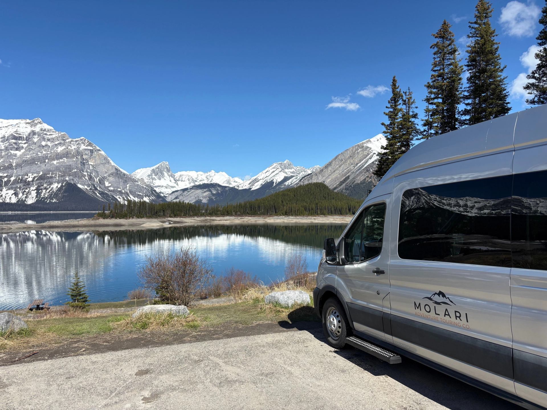 Silver Molari tour van parked beside calm mountain lake with snowy peaks reflected.