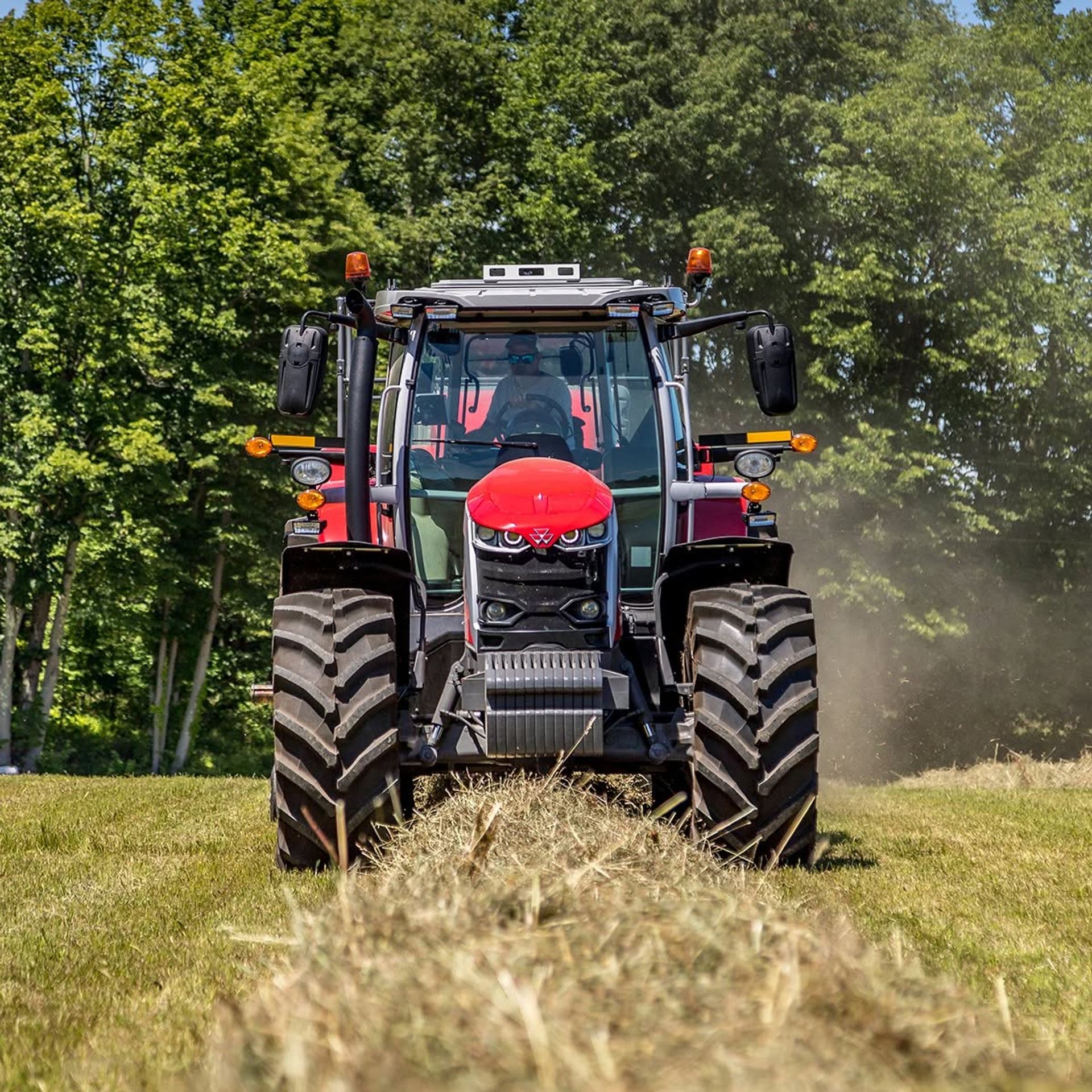 Red tractor drives over cut hay in a field bordered by tall green trees.