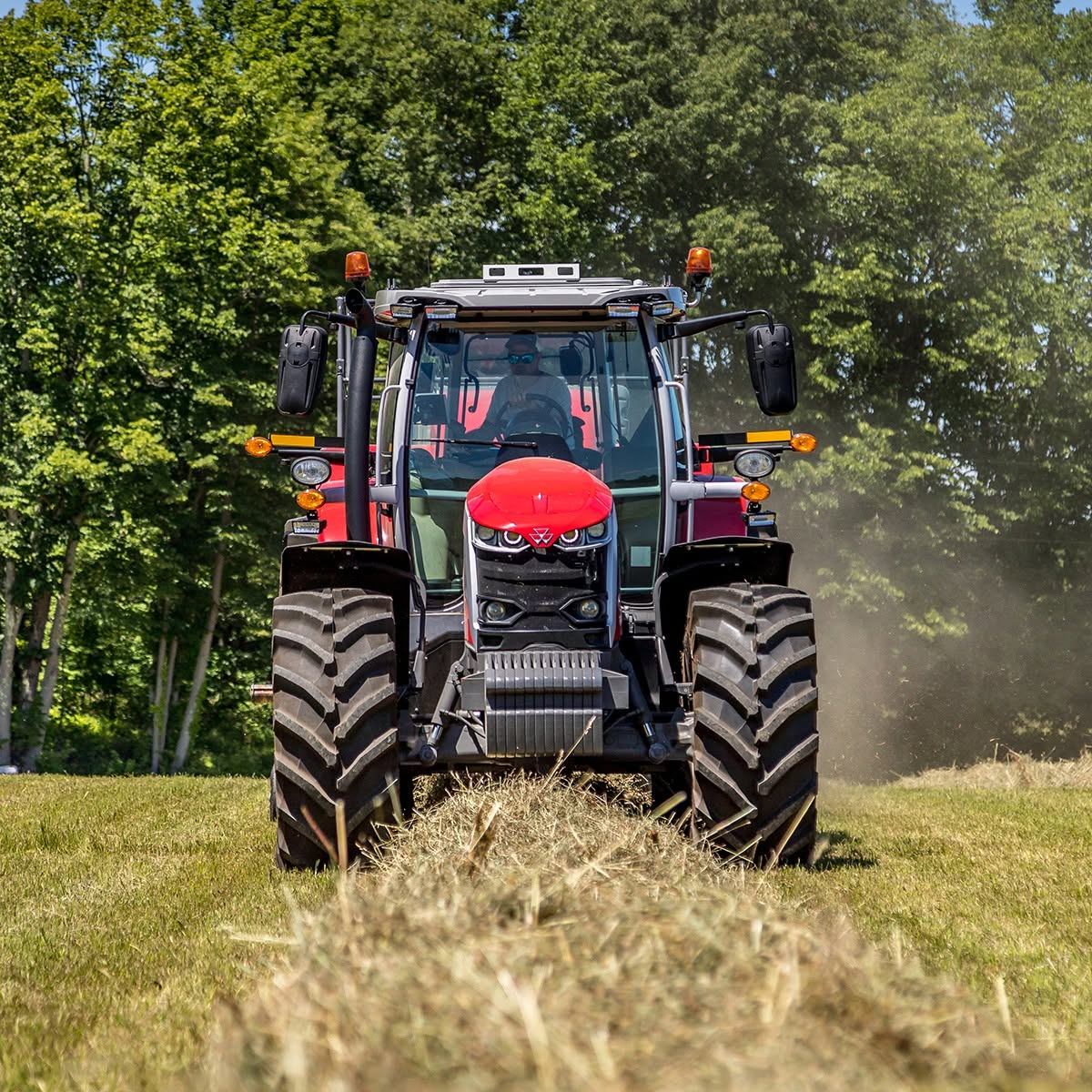 Red tractor drives over cut hay in a field bordered by tall green trees.